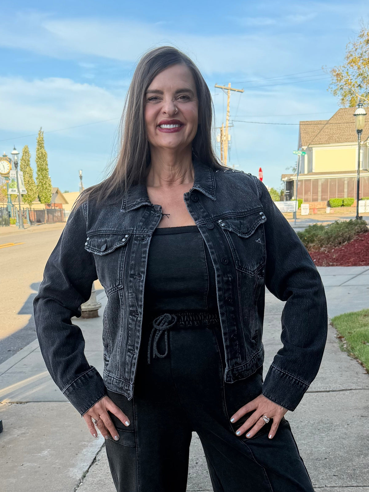 A woman stands on a sidewalk outdoors wearing a black denim jacket, black top, and black wide-leg pants. The background shows a street, grass, trees, and a blue sky.