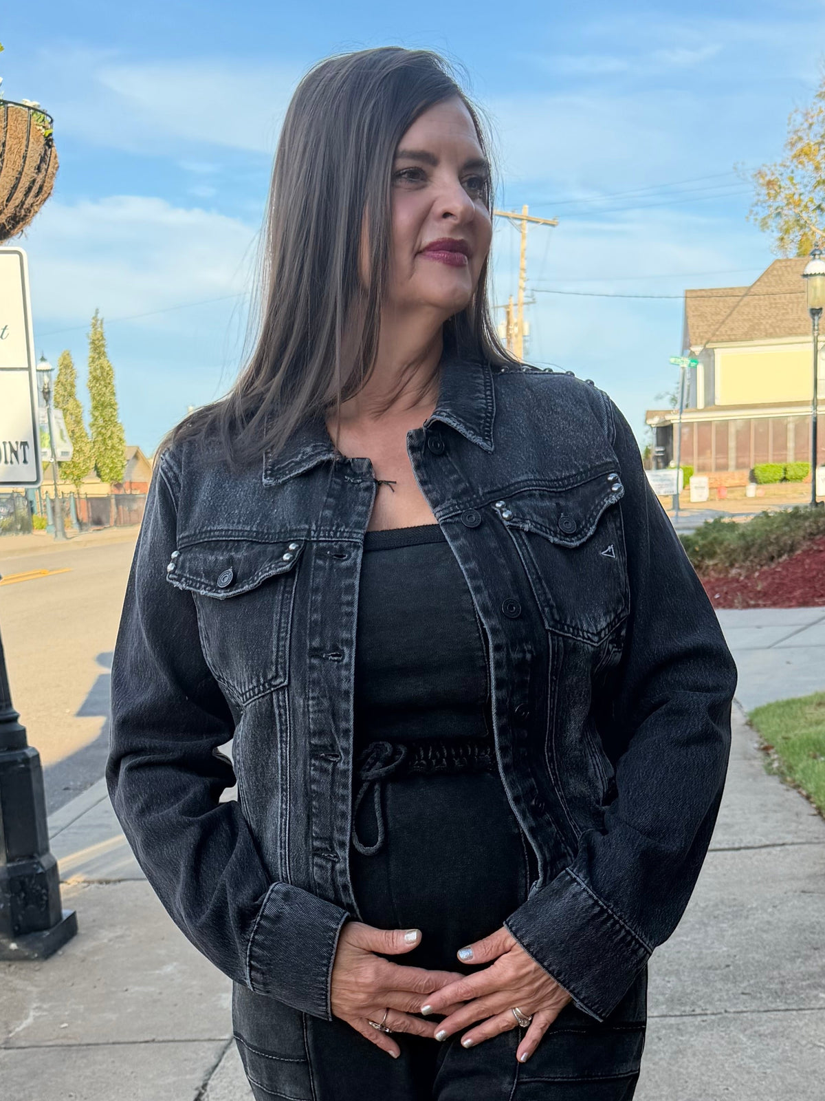 A woman stands on a sidewalk outdoors wearing a black denim jacket, black top, and black wide-leg pants. The background shows a street, grass, trees, and a house under a blue sky.