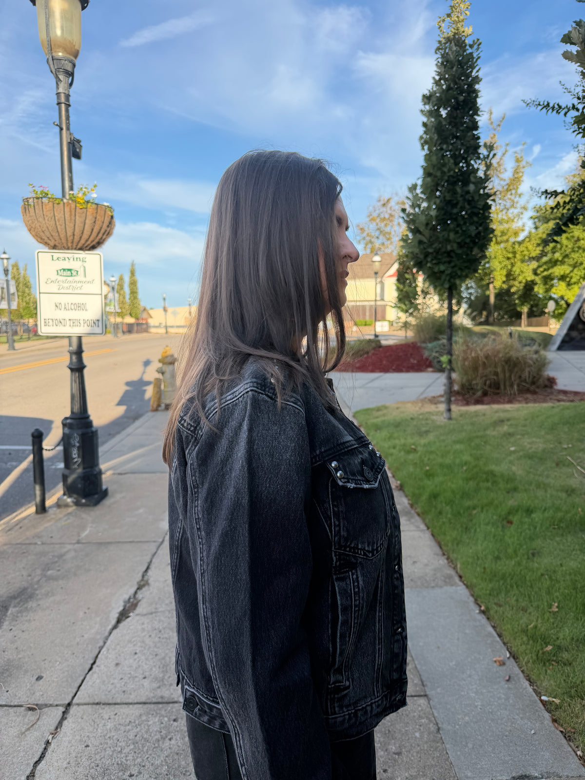 A woman with long dark hair stands in profile on a sidewalk outdoors, wearing a dark washed denim jacket. The background shows a street, grass, trees, and a blue sky.