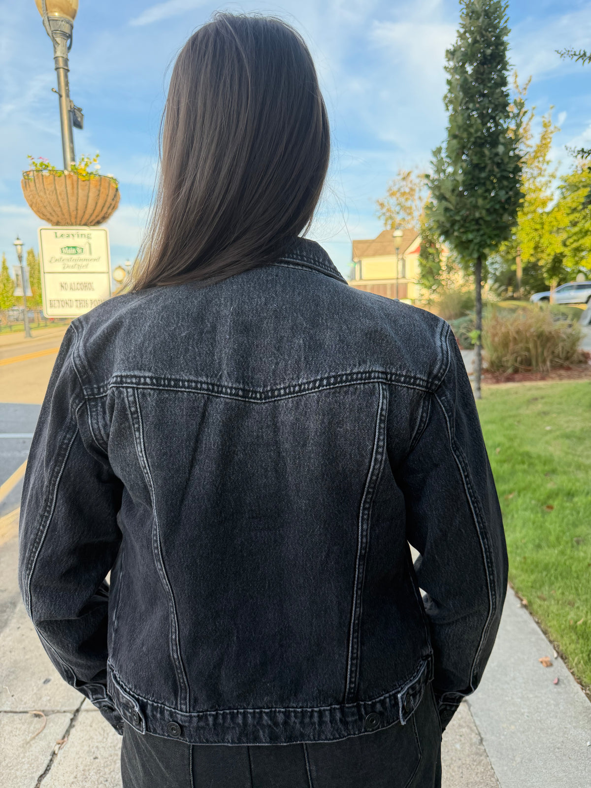 A person viewed from behind wearing a dark washed denim jacket outdoors on a sidewalk, with trees, grass, and a street sign in the background.