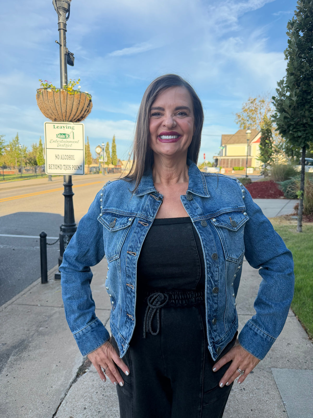 A woman smiling outdoors on a sidewalk, wearing a blue denim jacket over a black top and black pants. A street sign and lamp post are visible behind her, along with trees, grass, and houses under a blue sky.