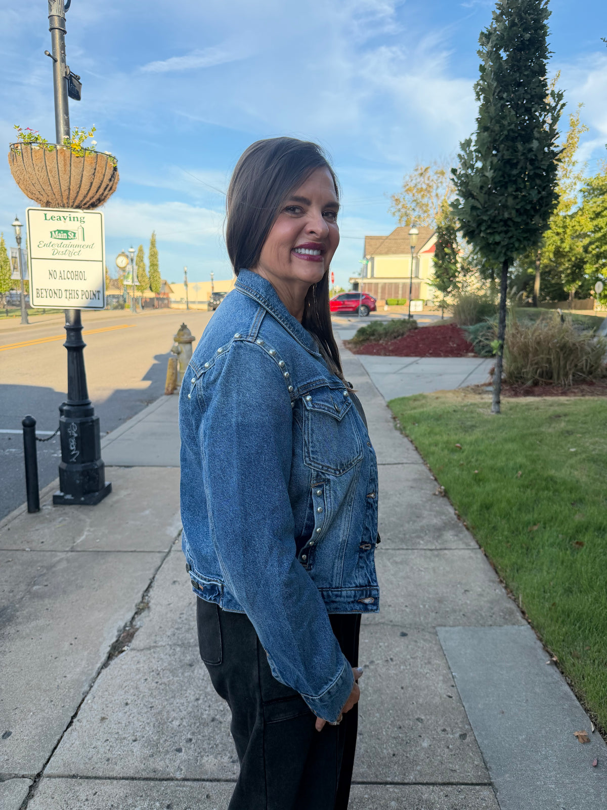 A woman wearing a blue denim jacket with silver studs and black pants, standing on a sidewalk outdoors.