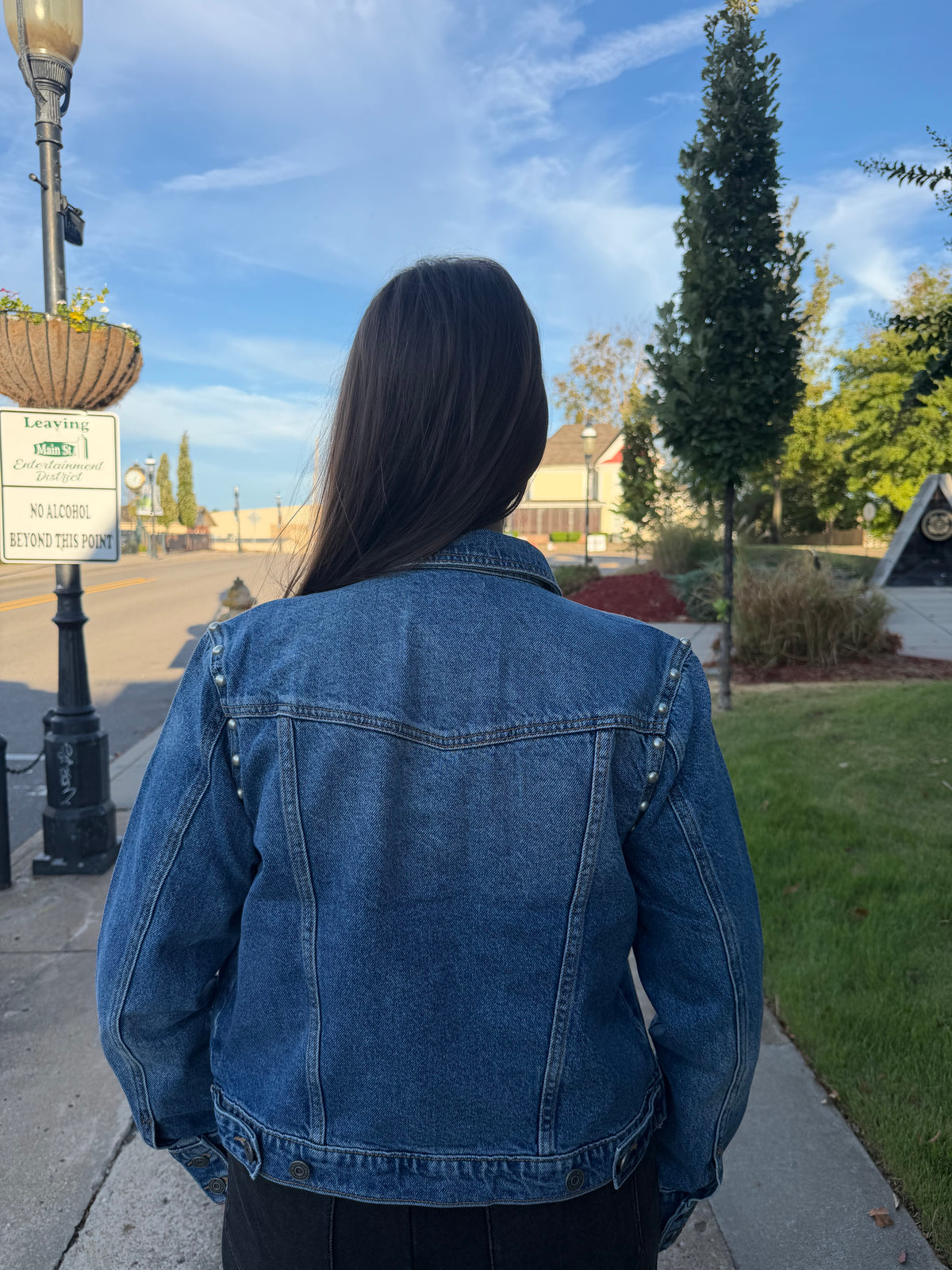A person viewed from behind wearing a blue denim jacket outdoors on a sidewalk, with trees, grass, a street, and a lamp post in the background.