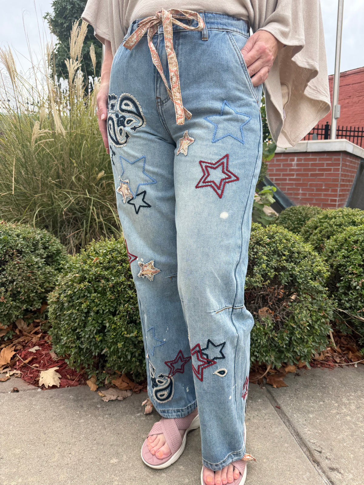 Light blue denim jeans with embroidered star patches and a patterned drawstring waist, shown on a model outdoors on a sidewalk.
