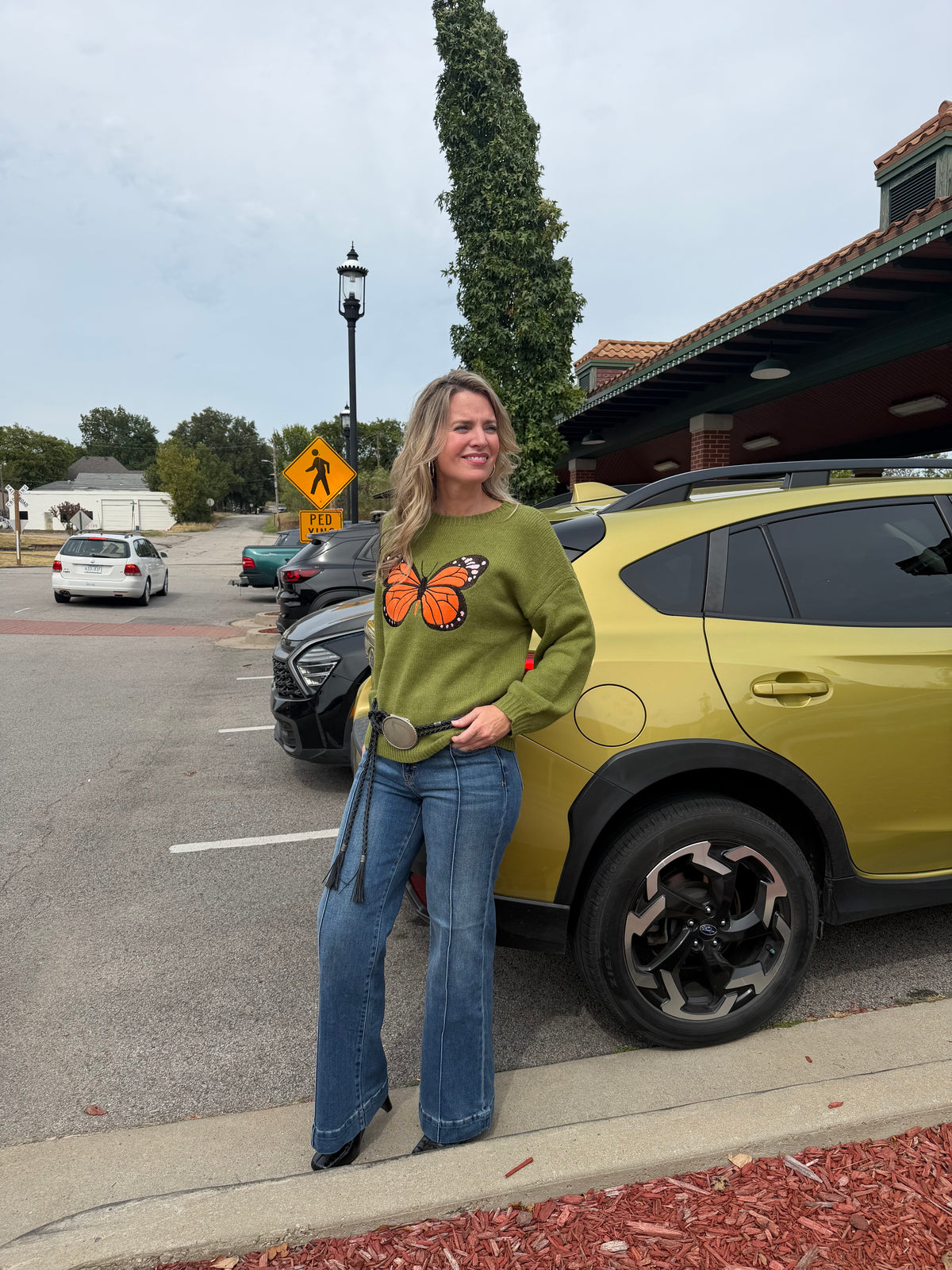 A woman stands outdoors in a parking lot next to a olive green SUV, wearing an olive green long-sleeve sweater with a large orange and black butterfly graphic on the front, blue jeans, and a large silver belt buckle.