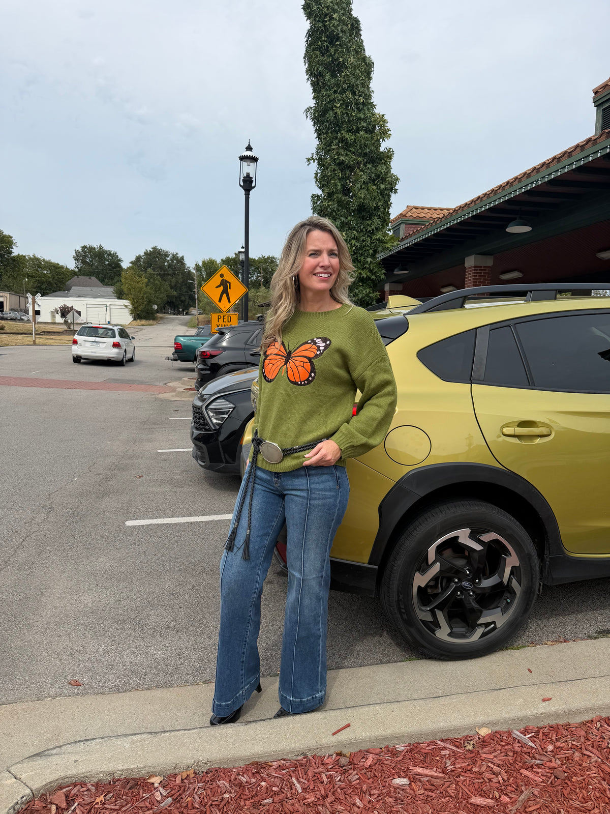 A woman stands in a parking lot wearing a green long-sleeve top with a large orange and black butterfly graphic on the front, paired with blue jeans. A yellow car is parked next to her.