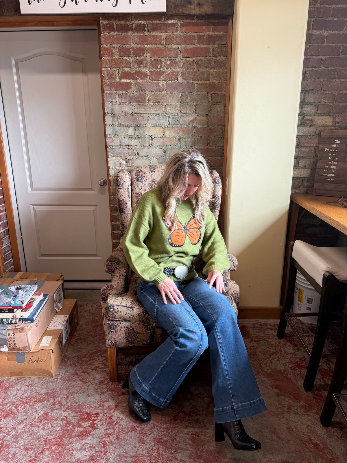A woman sits in a vintage upholstered armchair in a rustic room with brick walls, wearing a green crewneck sweater with a butterfly graphic, blue flare jeans, and black ankle boots.