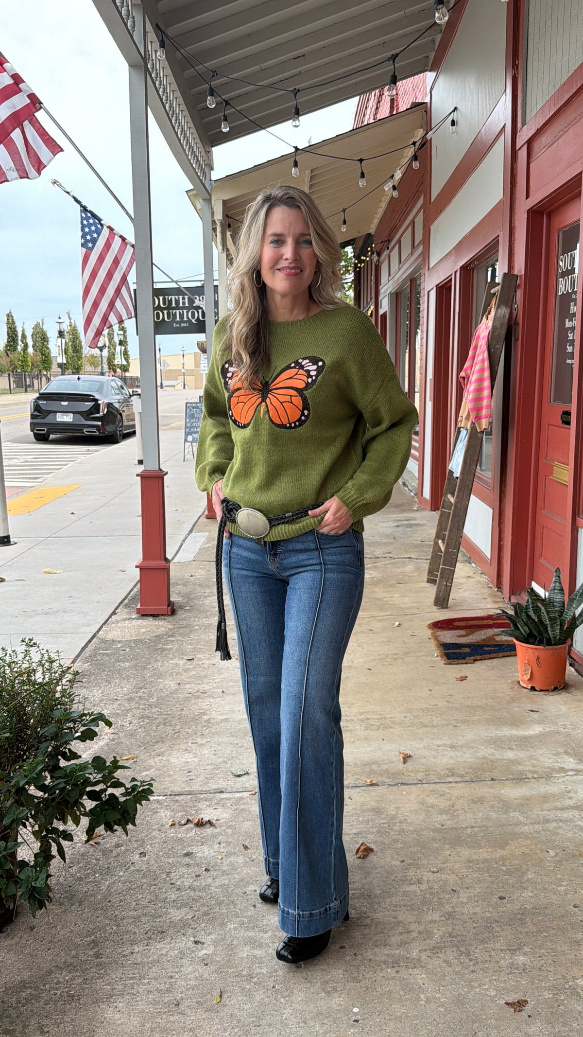 A woman stands on a storefront sidewalk wearing an olive green sweatshirt with a large orange and black butterfly graphic on the front, paired with blue jeans and black shoes.