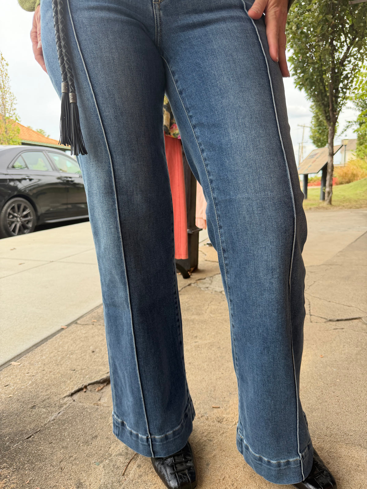 A close-up of a person wearing blue denim jeans with a distinctive vertical seam running down the front of each leg and a black tassel detail at the waistband, standing on a sidewalk outdoors.