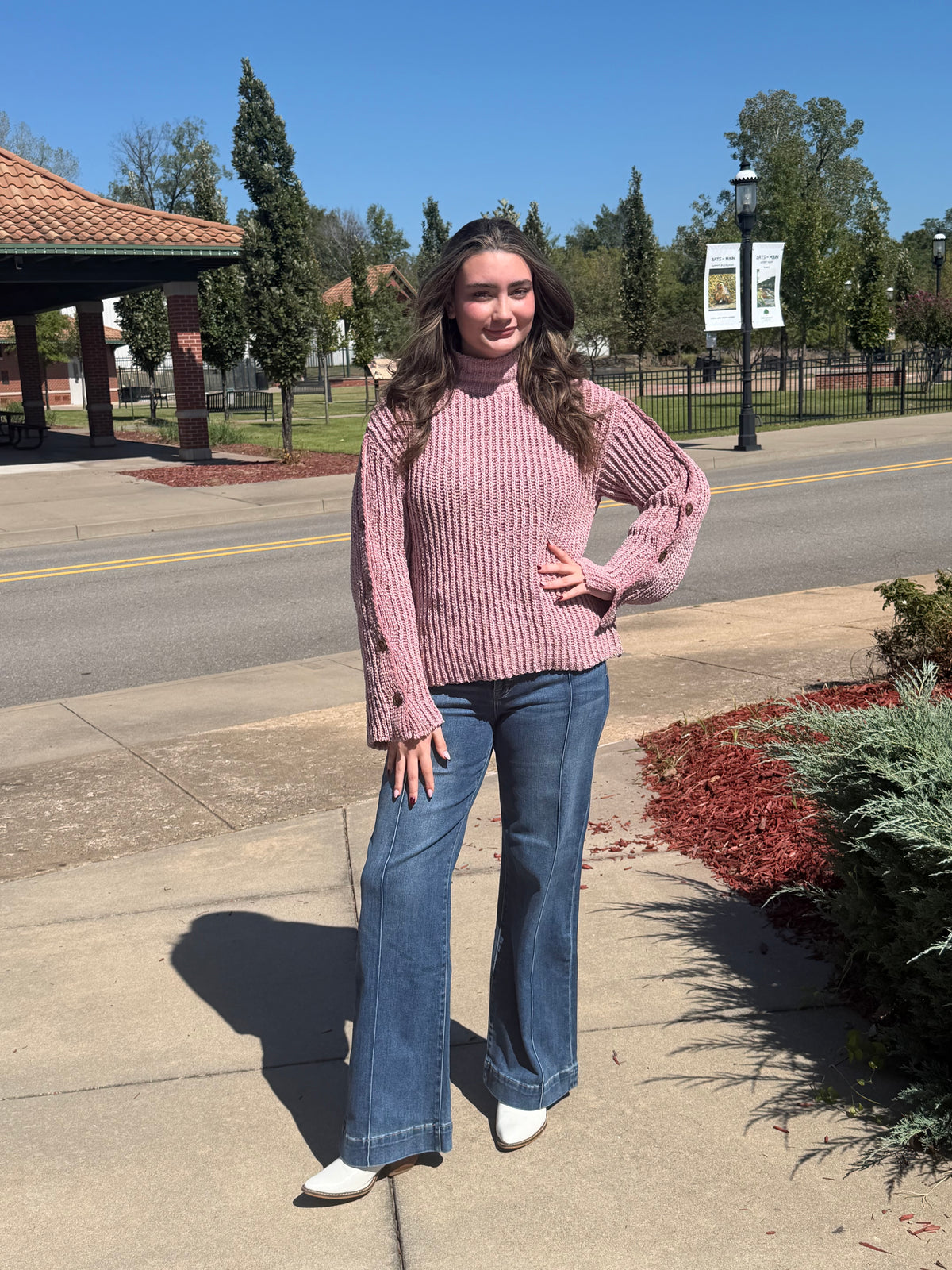 Front view of a model wearing a dusty mauve ribbed sweater with button-detail sleeves, wide-leg jeans, and white boots outdoors.
