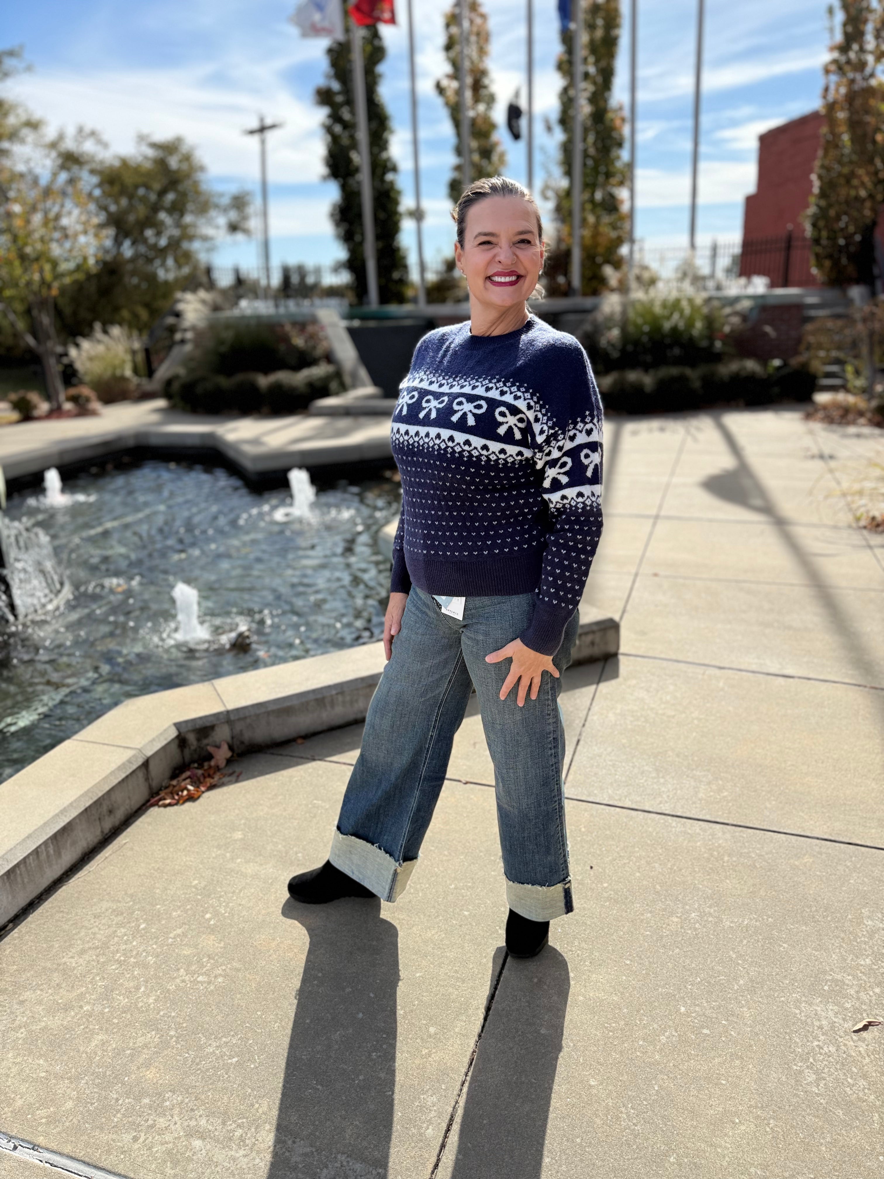 Person wearing a navy sweater with white patterns and jeans standing outdoors near a water feature.