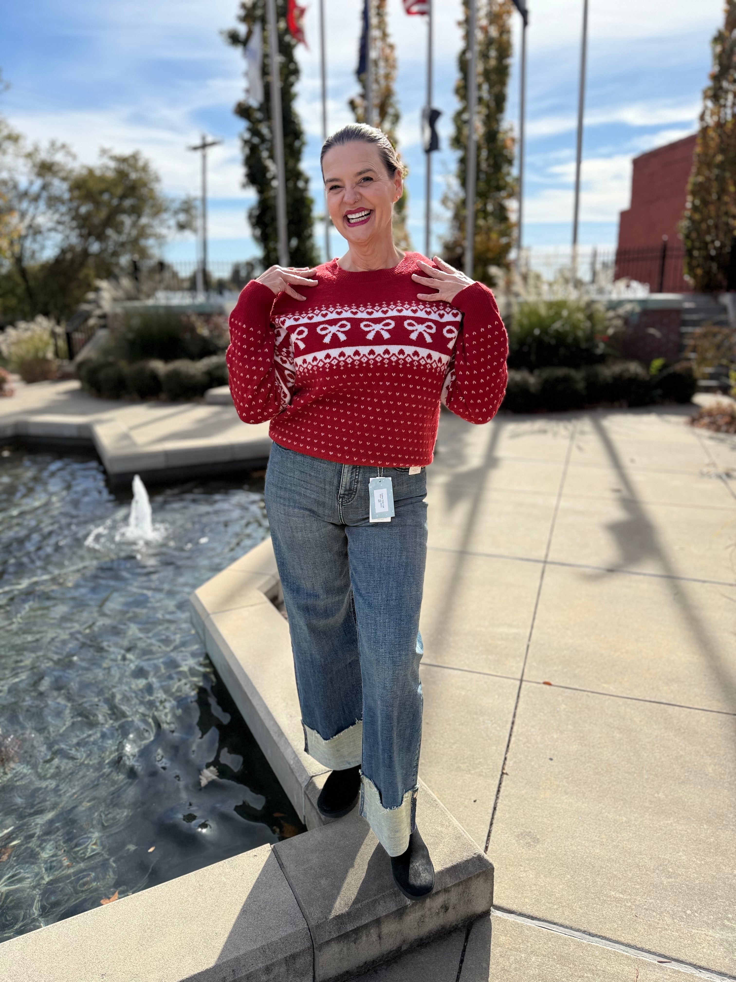 Woman wearing a red sweater with white patterns and blue jeans standing by a water fountain.