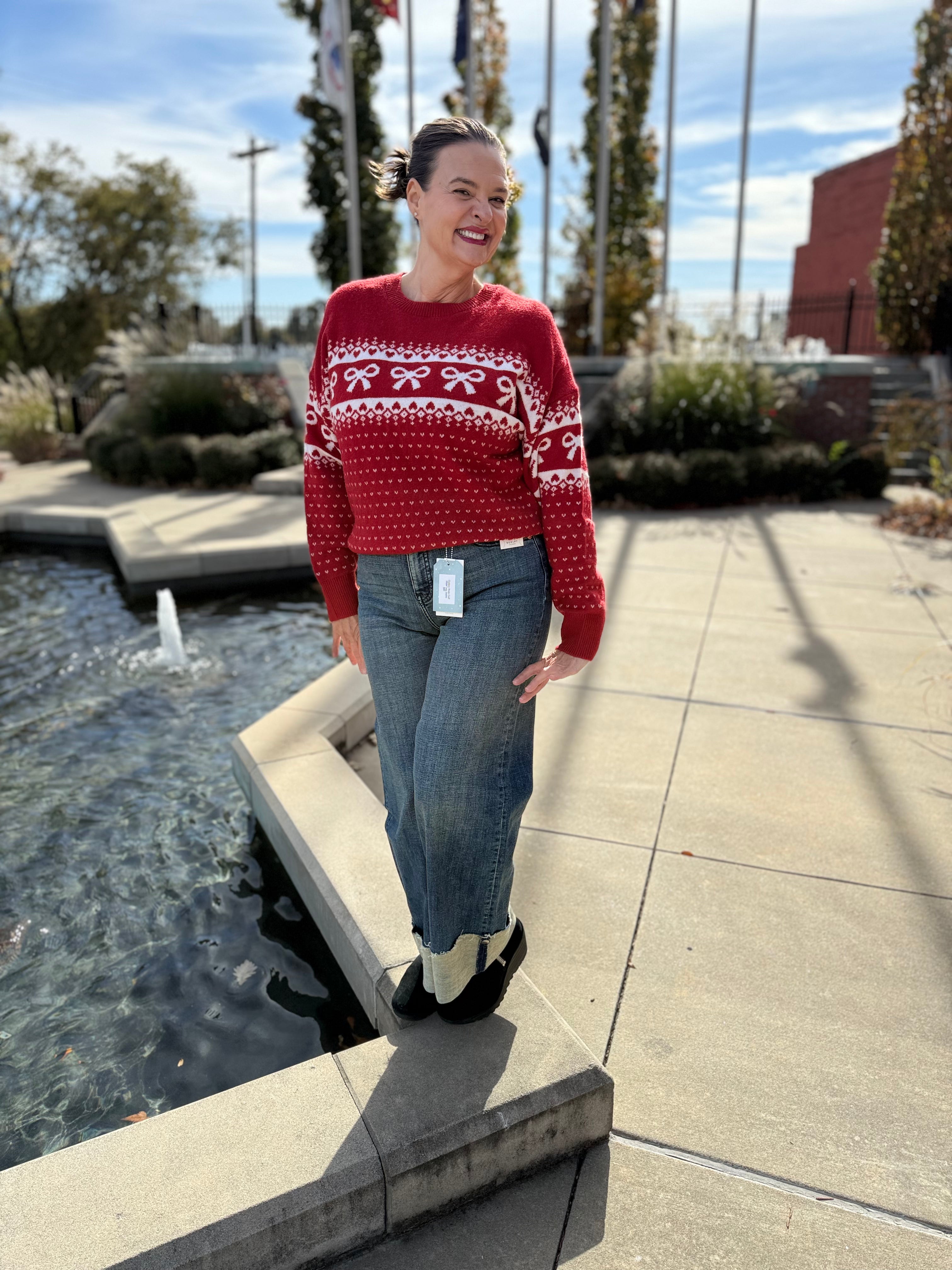 Woman wearing a red sweater with white patterns and blue jeans standing by a water fountain.