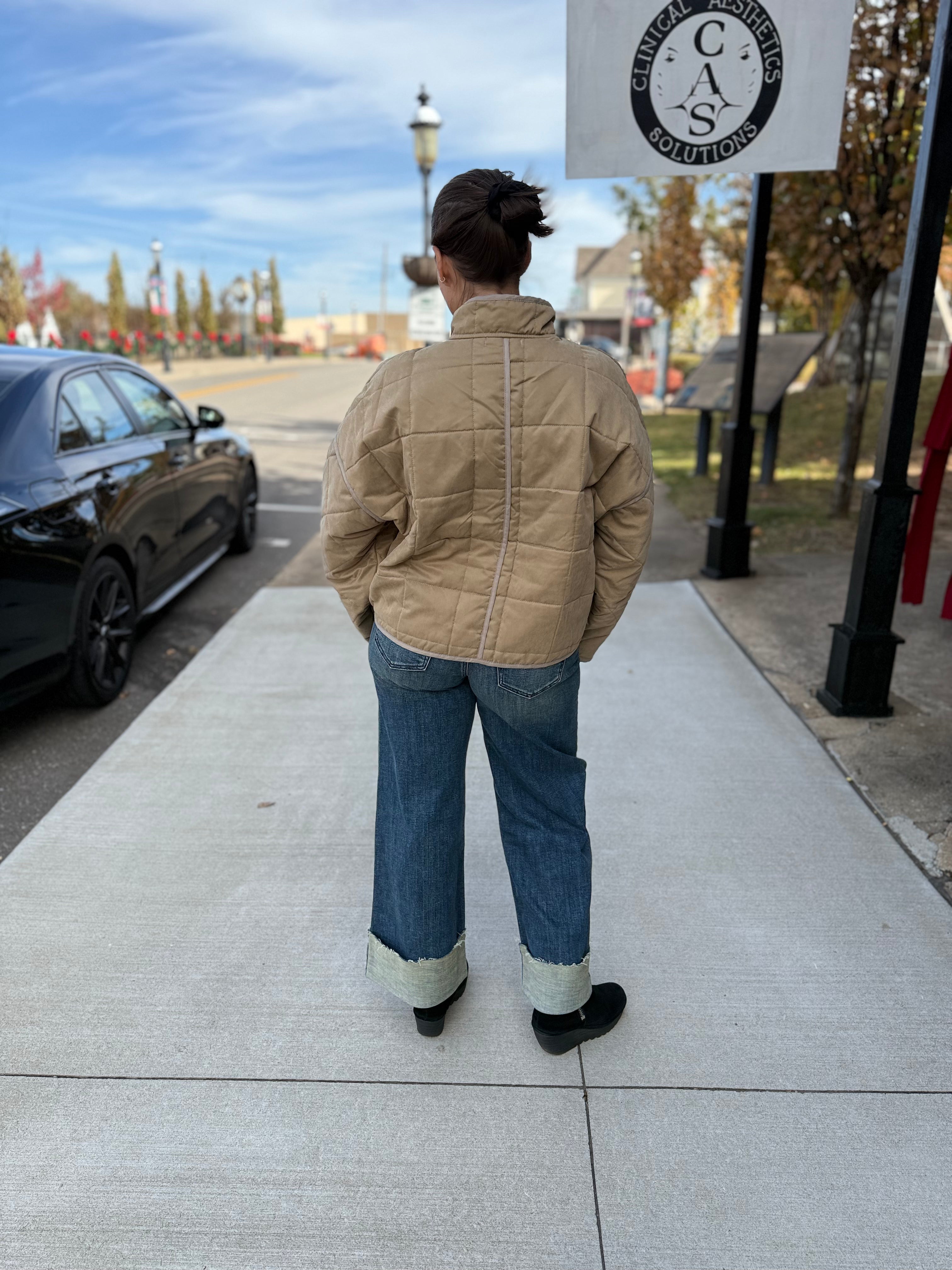 Person wearing a beige jacket and blue jeans standing on a sidewalk with a street sign in the background.