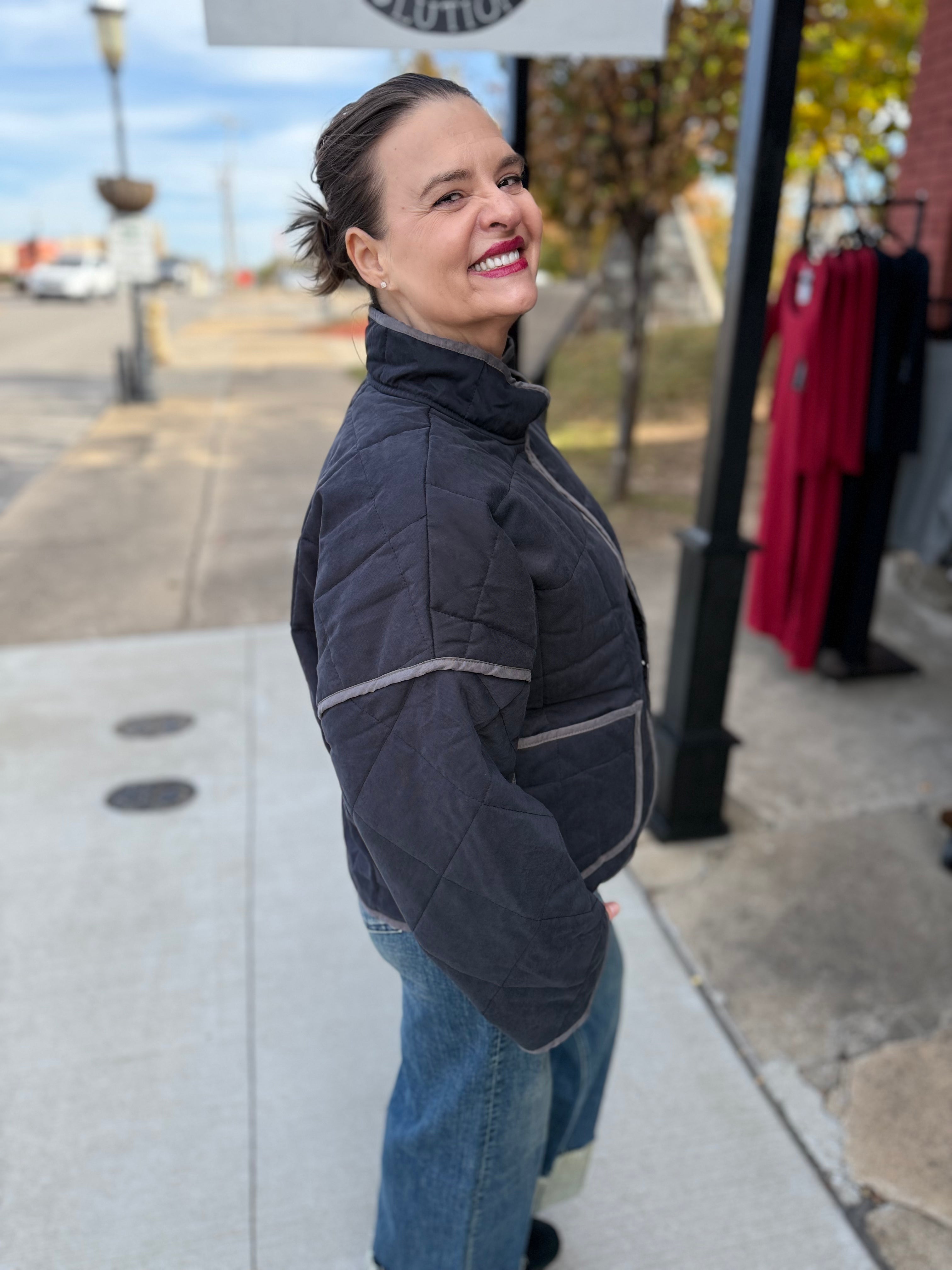 Woman wearing a dark jacket and jeans standing on a sidewalk with a store display in the background.