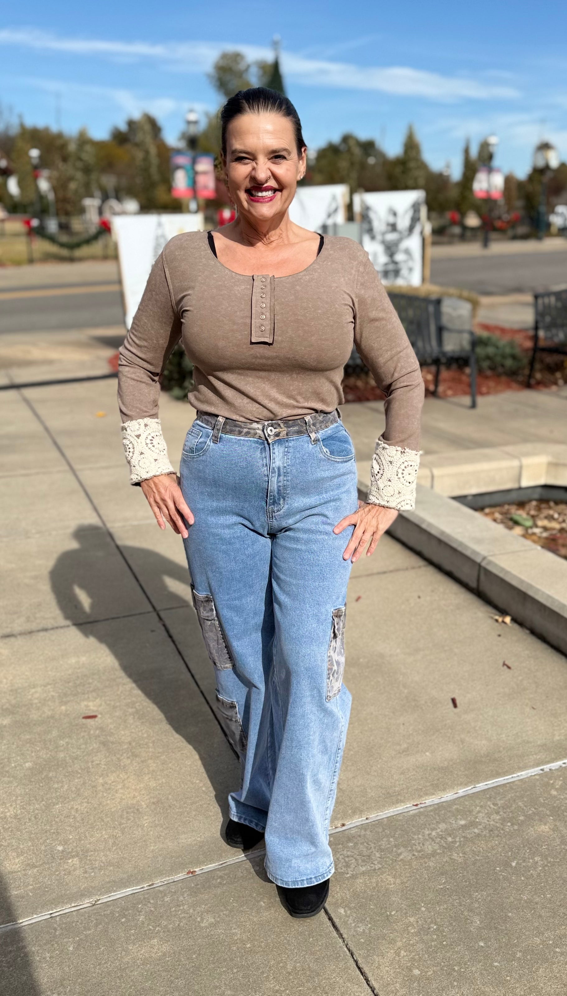 Woman wearing a brown top and blue jeans standing on a sidewalk with trees and buildings in the background.