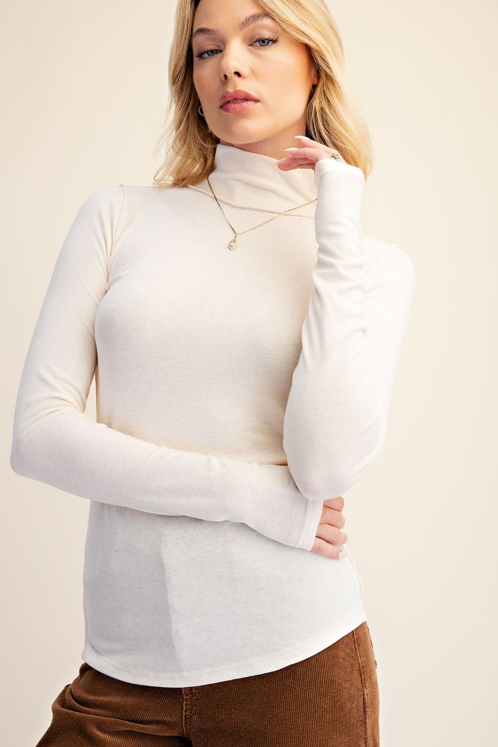 A woman wearing a fitted, long-sleeve mock neck turtleneck top in a warm off-white or cream color, paired with brown pants, against a solid light beige background.