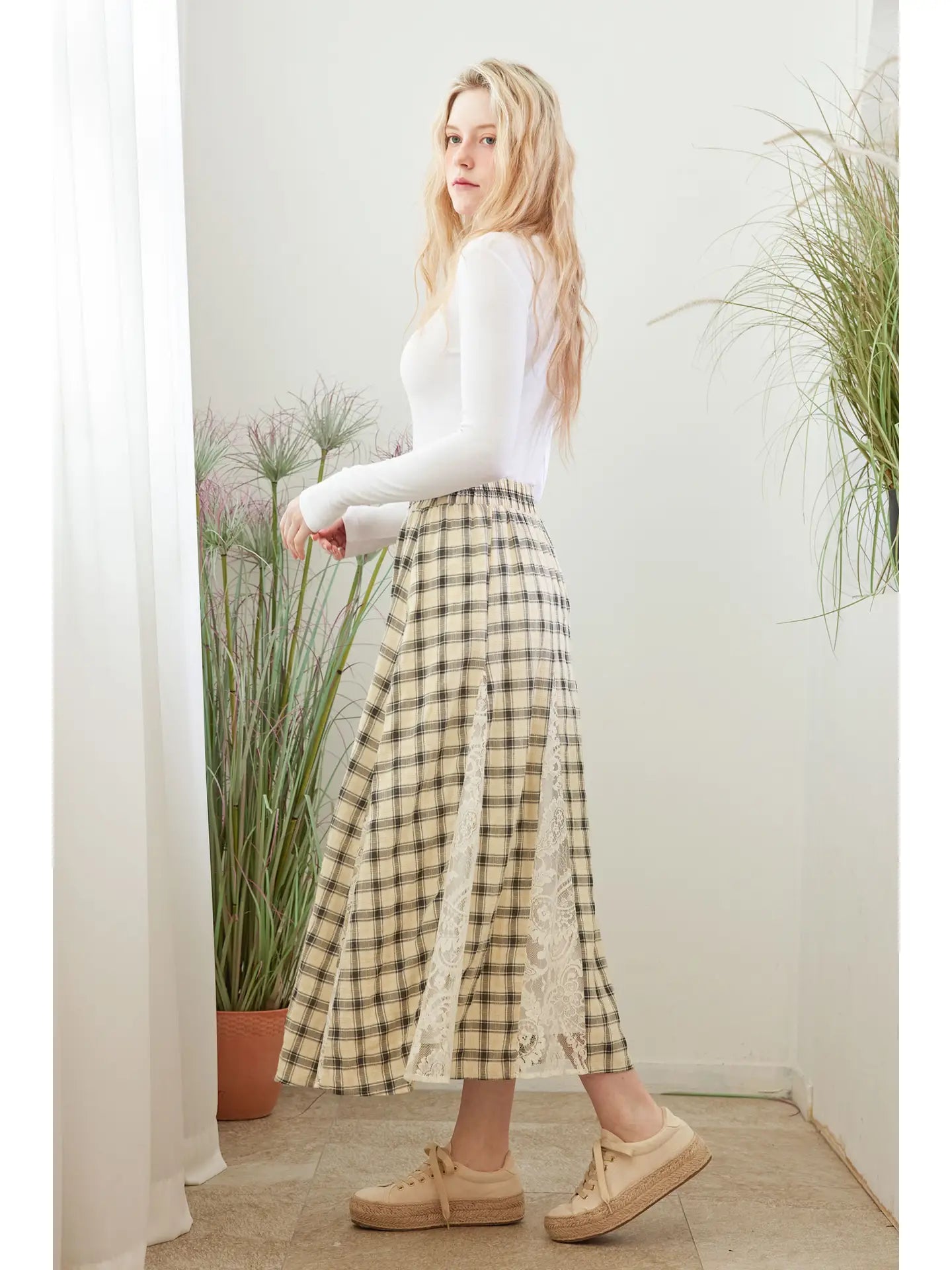 Woman wearing a white long-sleeve top and plaid skirt standing indoors with plants in the background.
