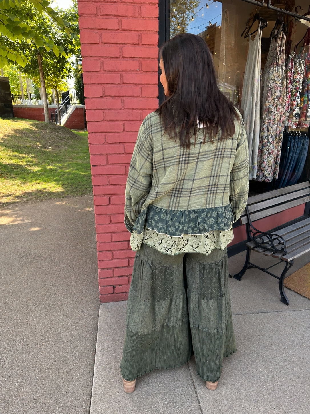 A woman stands with her back to the camera wearing a loose, vintage-style plaid shirt and wide-leg skirt set in olive green tones, posing outdoors next to a red brick pillar and a clothing boutique window.