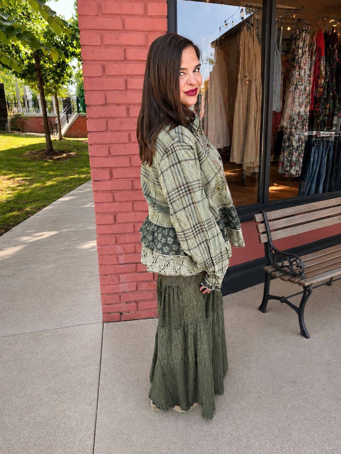 A woman stands outdoors in front of a storefront, wearing a loose sage green plaid jacket with lace trim over a long olive green skirt. A brick pillar, concrete walkway, bench, and clothing store window are visible in the background.