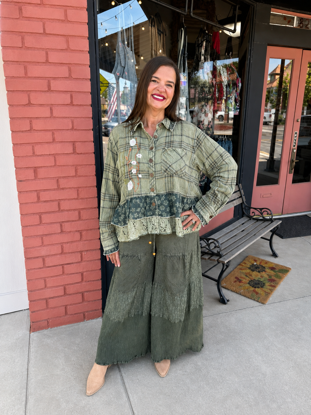 A woman stands on a sidewalk in front of a storefront, wearing a long-sleeve button-front blouse with a green plaid and lace ruffle hem, paired with wide-leg olive green pants and beige pointed shoes.