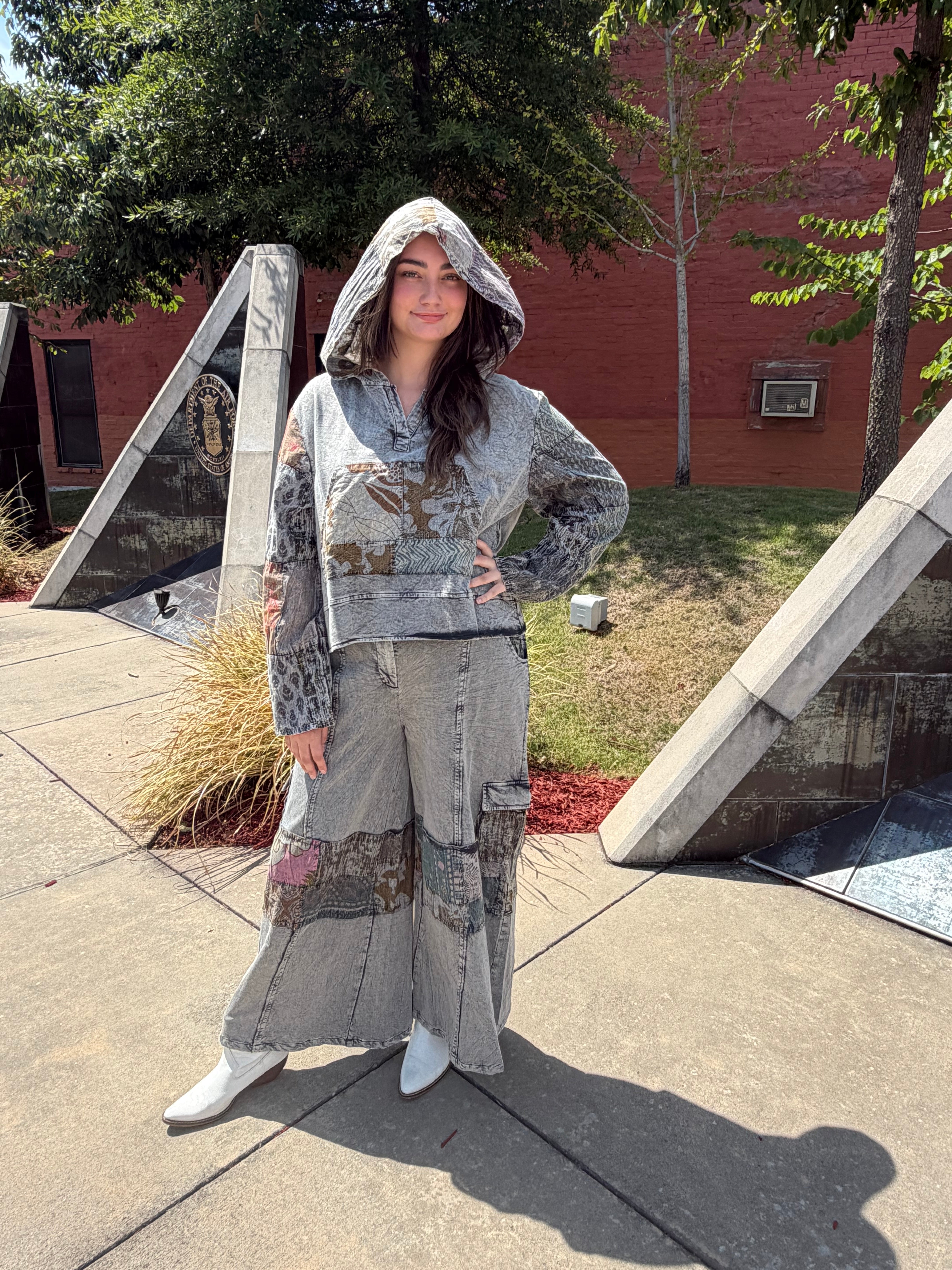 A woman stands outdoors on a concrete plaza wearing a matching gray patchwork hoodie and wide-leg patchwork pants with white boots. Trees, a brick building, and geometric concrete sculptures are visible in the background.