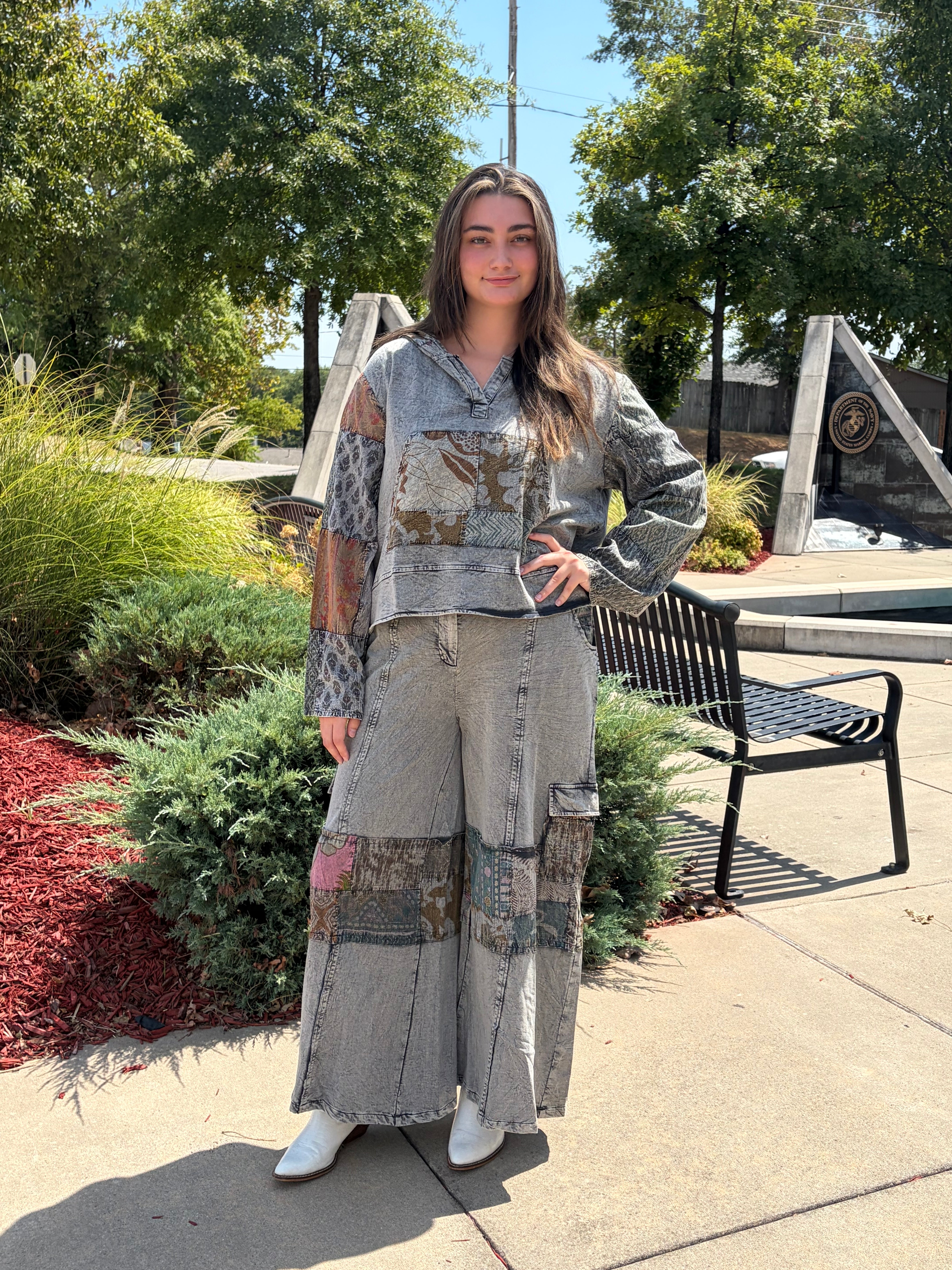 A woman stands outdoors on a paved walkway wearing a matching patchwork denim outfit consisting of a cropped long-sleeve top and wide-leg pants, with white boots. Trees, shrubs, and a bench are visible in the background.