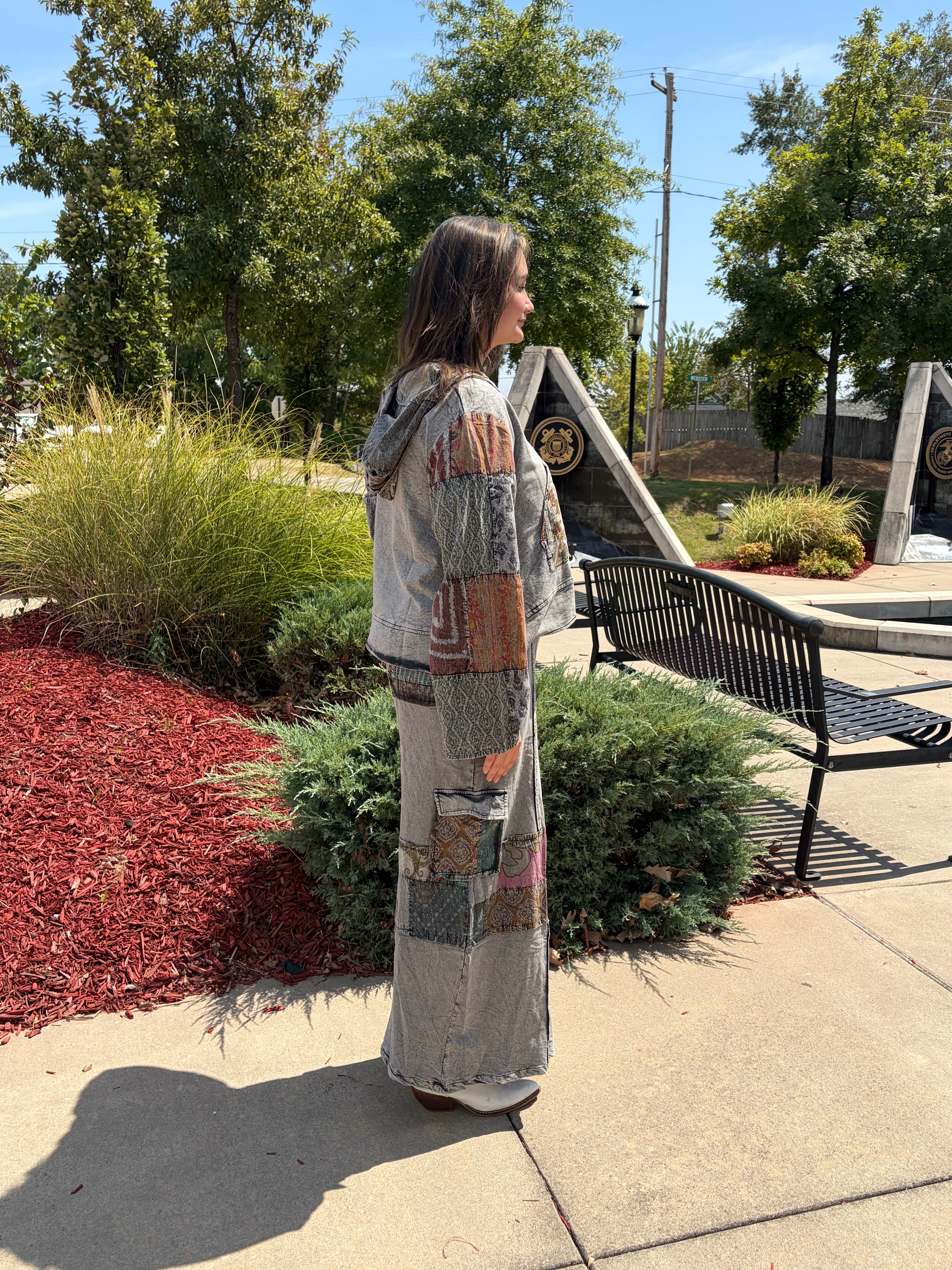 A woman stands outdoors in a park-like setting wearing a long, patchwork-patterned hooded garment. The garment has a mix of earthy tones including beige, brown, gray, and muted orange in a patchwork design.