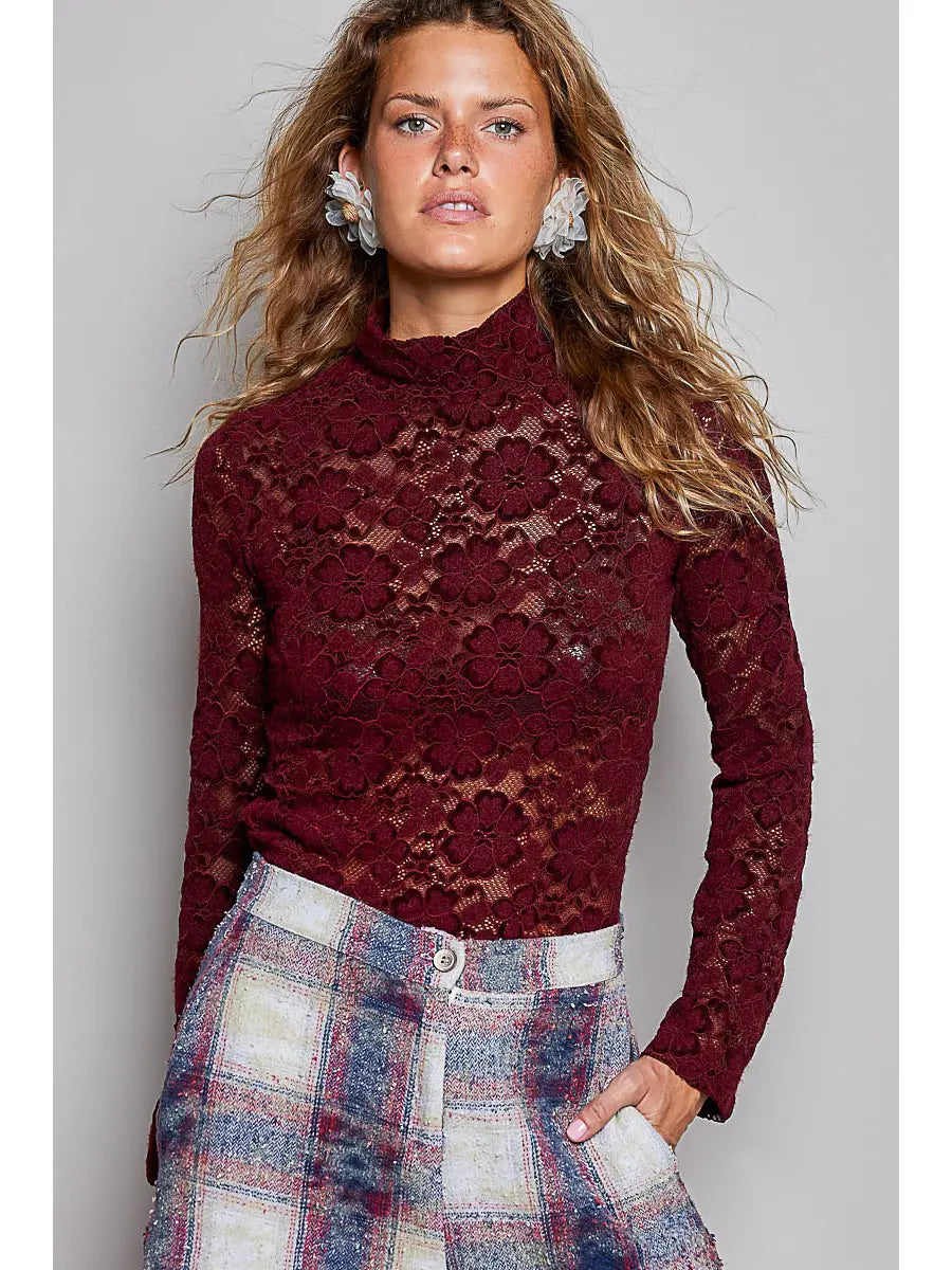 A woman wearing a burgundy lace mock-neck long-sleeve top and a blue, red, and white plaid skirt, standing against a solid gray studio background.