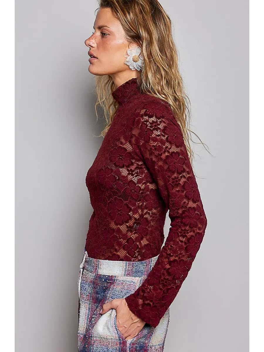 A woman shown in side profile wearing a long-sleeve burgundy lace mock-neck top and plaid pants against a plain gray studio background.