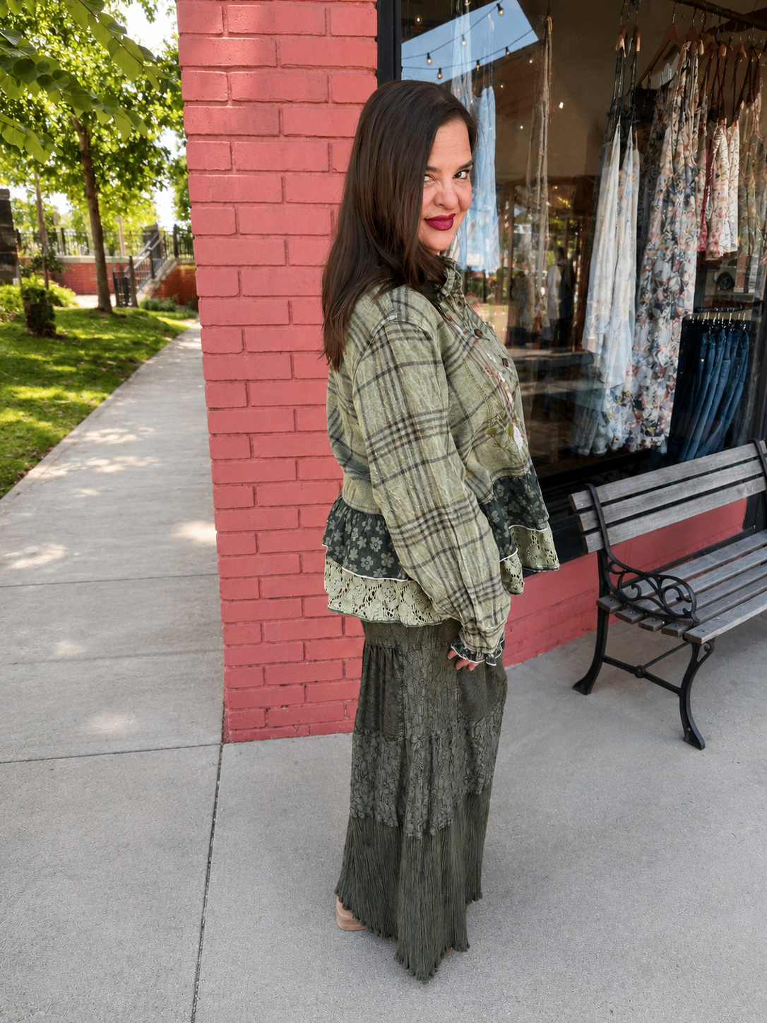 A woman stands on a sidewalk in front of a storefront, wearing a long-sleeve green plaid top with lace trim and a long dark green skirt. A pink brick pillar, a wooden bench, and a clothing window display are visible in the background.