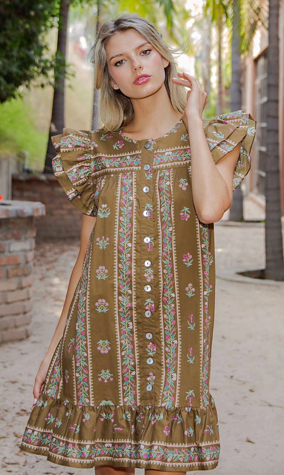 A woman wearing a brown bohemian-style dress with colorful floral embroidery and ruffled sleeves, standing outdoors on a sandy path with palm trees in the background.