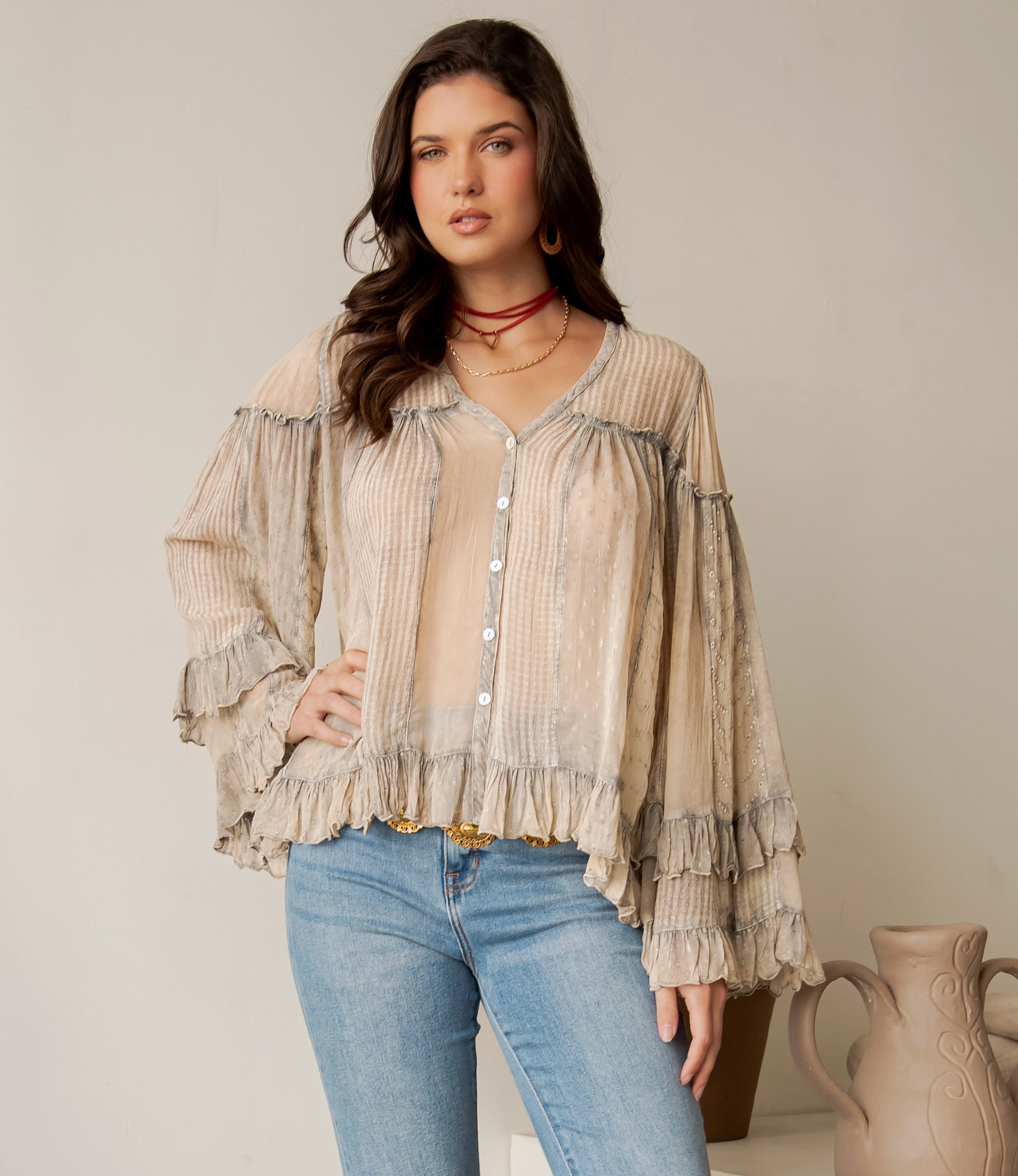 A woman models a beige and gray striped long-sleeve blouse with ruffles, paired with light blue jeans, standing against a neutral beige wall.