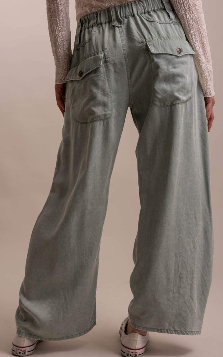 A person shown from the back wearing wide-leg sage green pants with an elastic waistband, buttoned back pockets, and a light beige lace-up top, standing against a plain beige studio background.