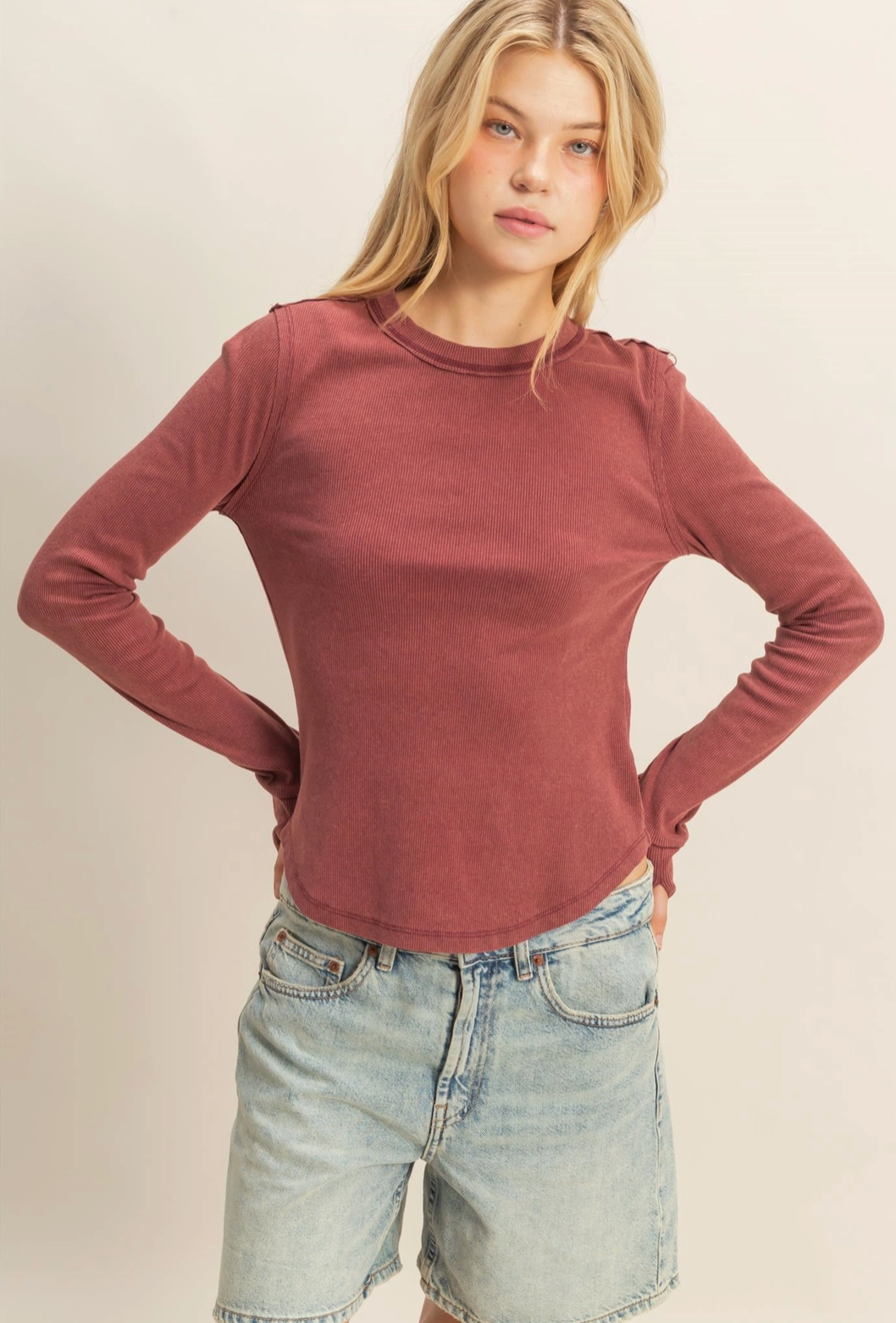 Woman wearing a maroon long-sleeve top and denim shorts on a beige background