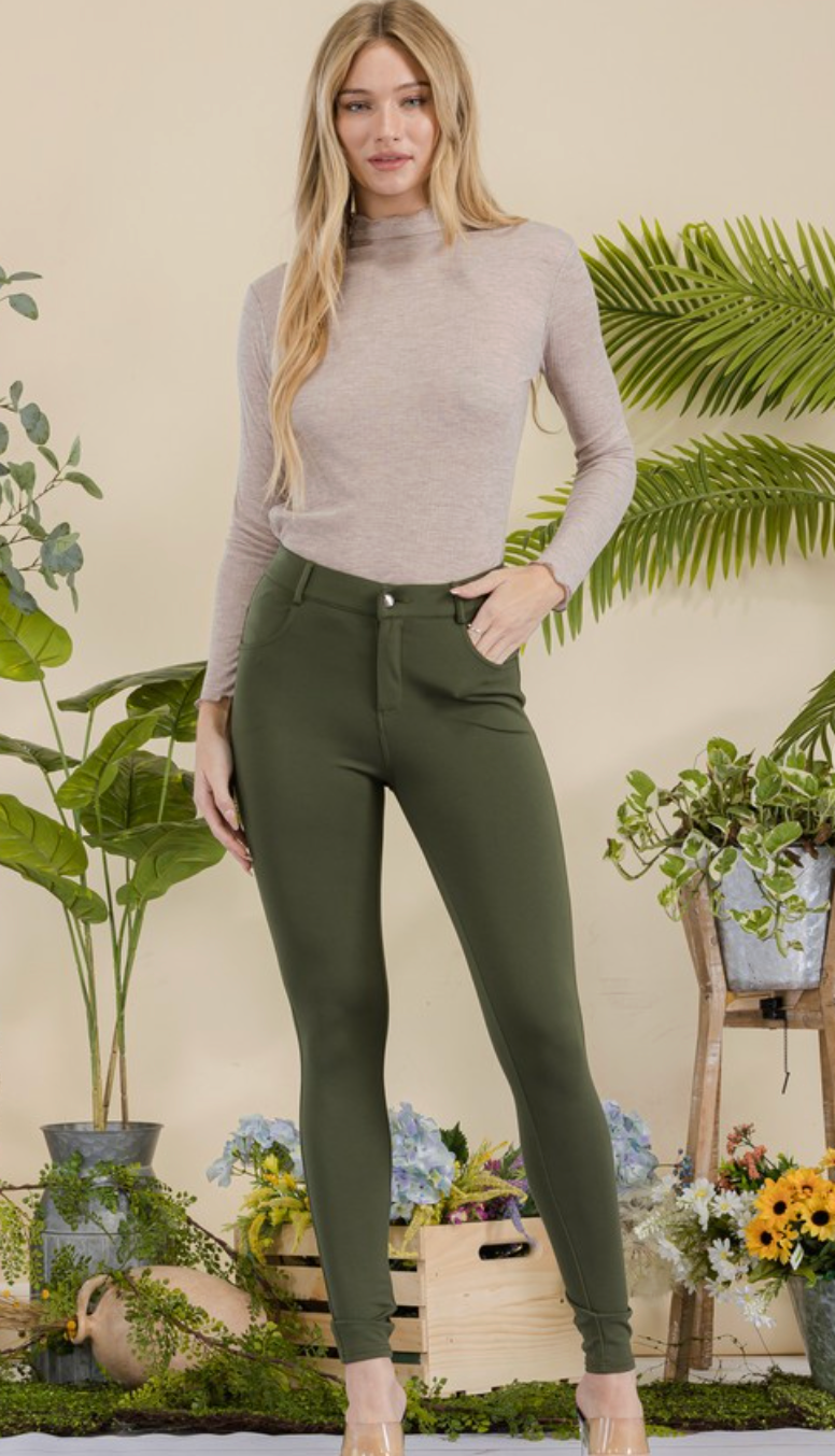 Woman wearing a beige long-sleeve top and green pants in a decorative setting with plants and flowers.