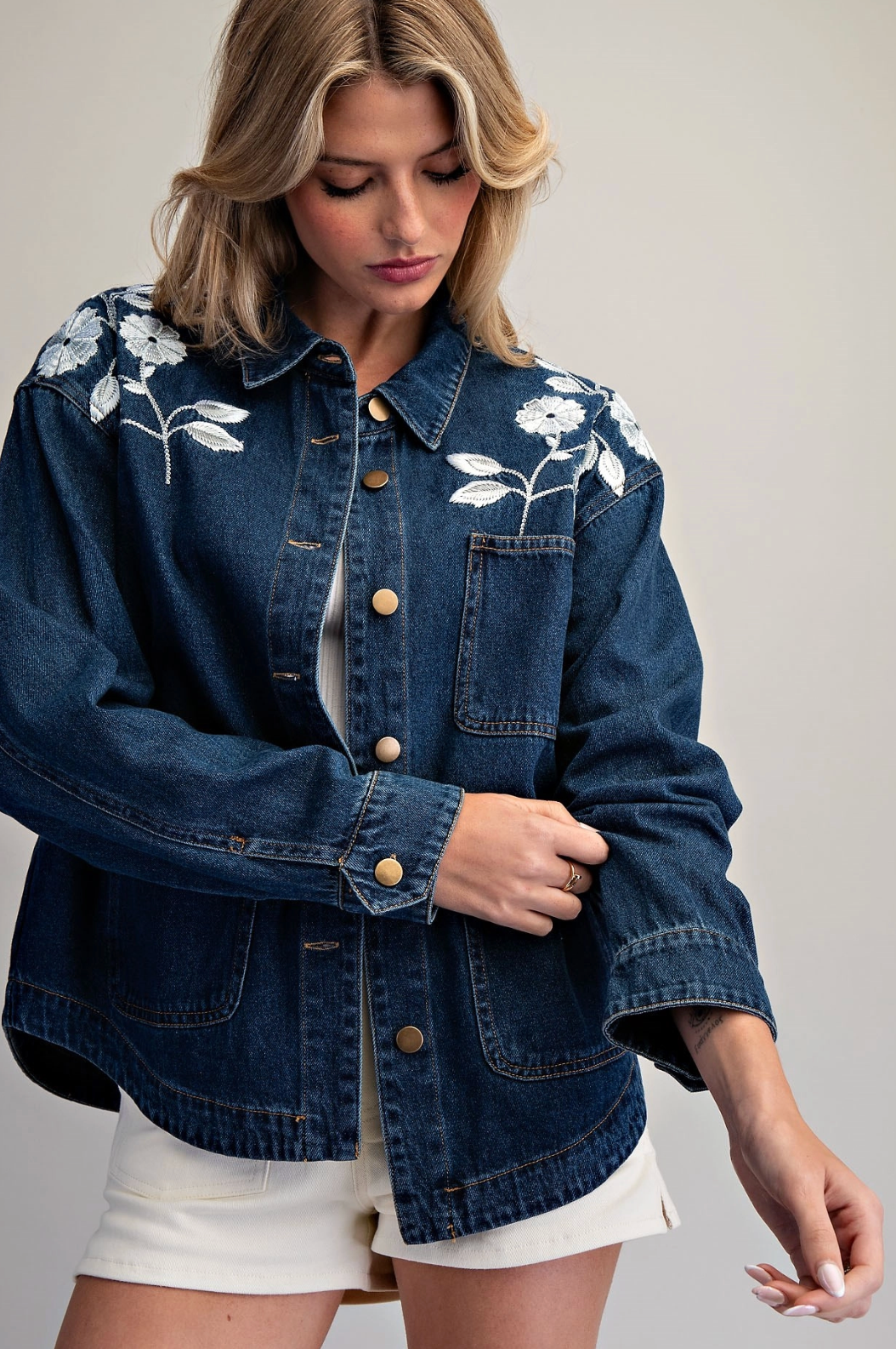 A woman wearing a dark blue denim jacket with white floral embroidery on the shoulders, styled with white shorts against a neutral studio background.