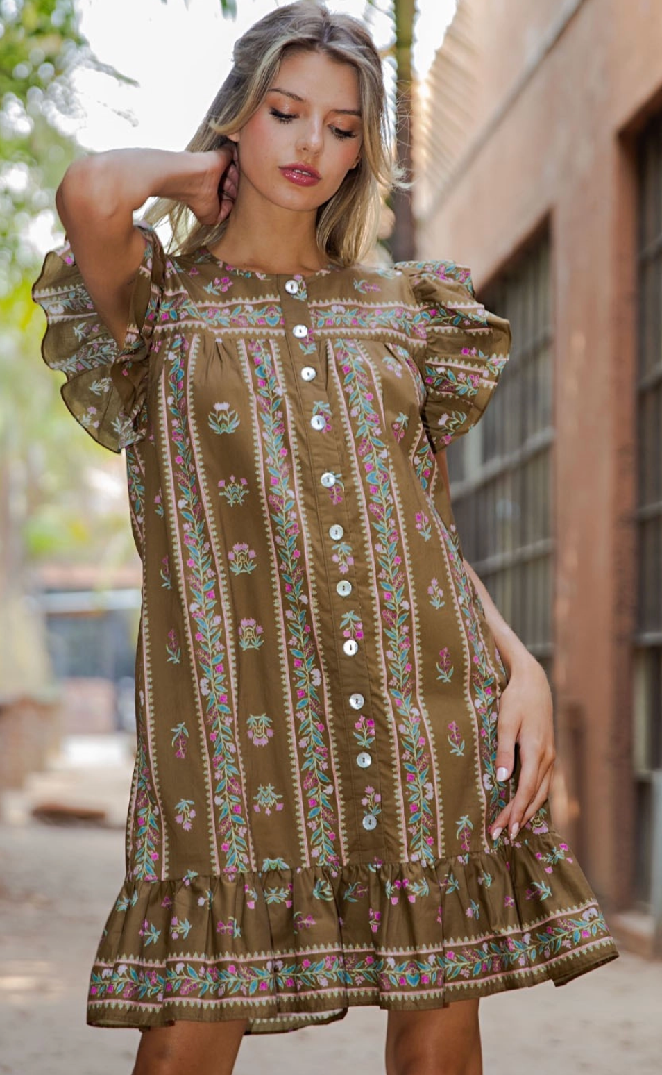 A woman wearing a brown bohemian-style mini dress with vertical floral embroidery and button details, posing outdoors against a blurred brick building background.