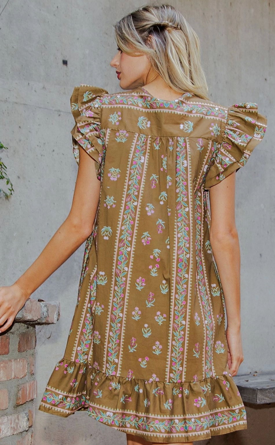 A woman wearing a brown bohemian-style dress with floral prints and ruffled sleeves, photographed from behind against a concrete wall.