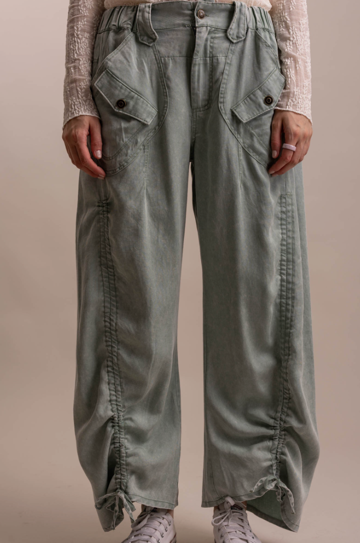A person wearing wide-leg sage green cargo pants with gathered side seams, flap pockets, and drawstring ankle cuffs, paired with a cream lace-trim top and white sneakers against a plain beige studio background.