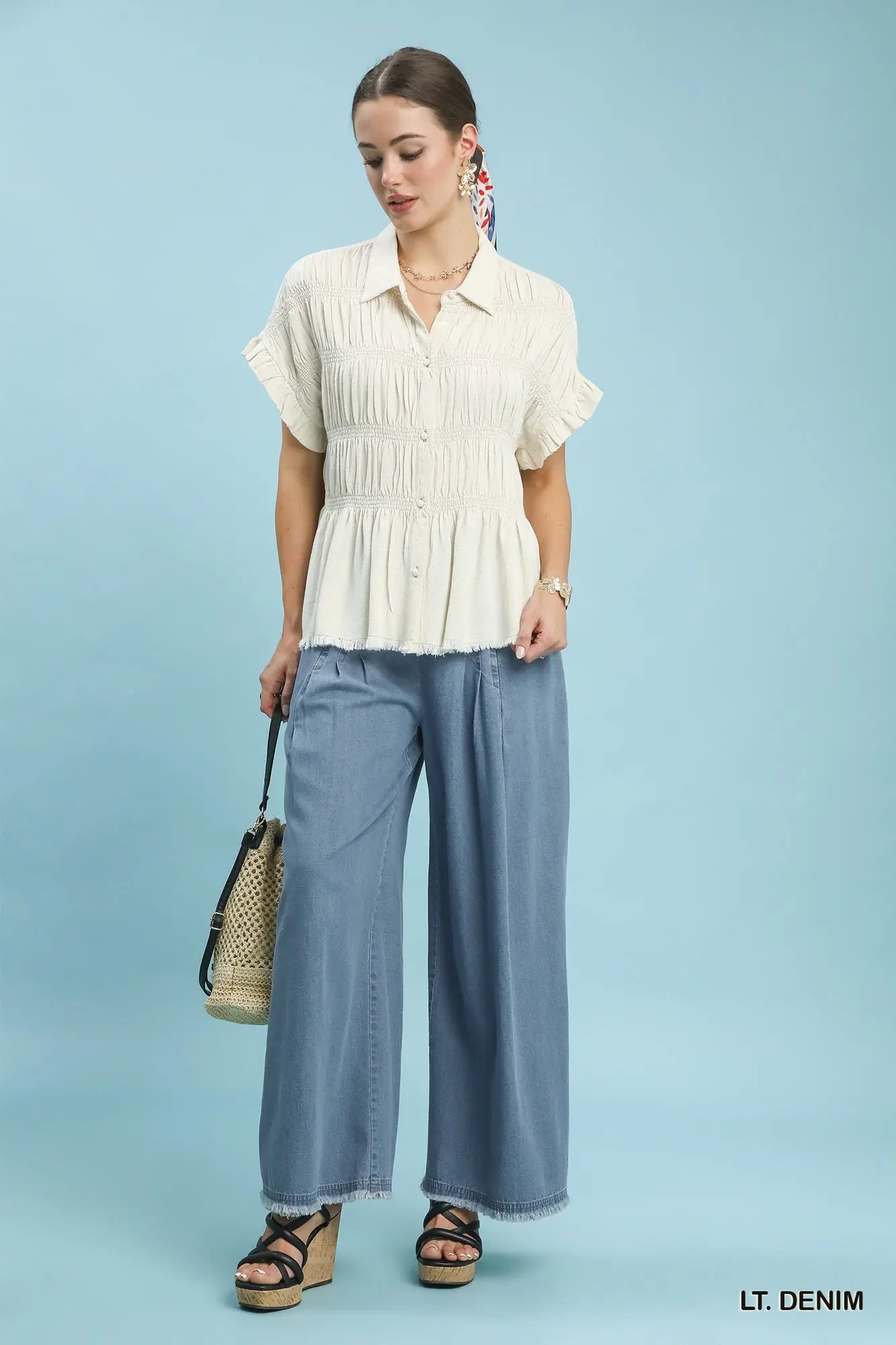 Woman wearing a white blouse and light blue pants holding a woven bag on a light blue background