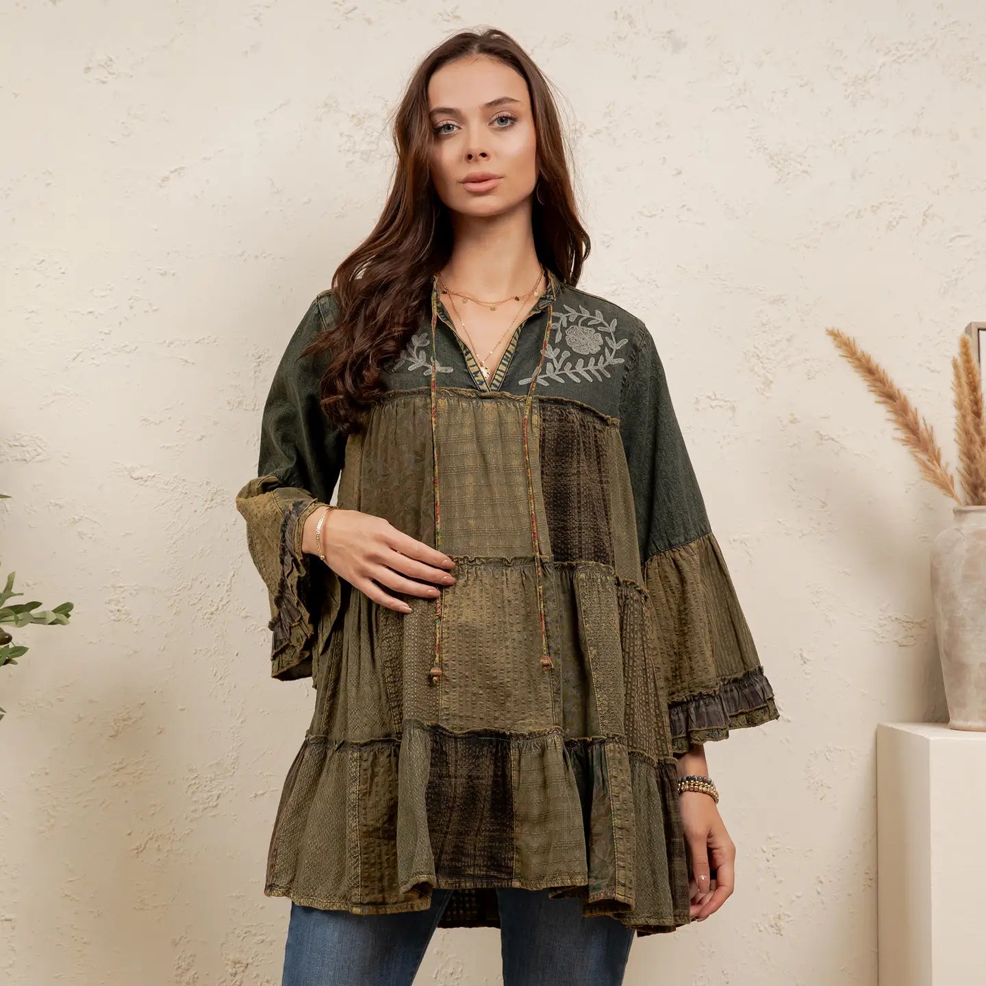 A woman models a tiered bohemian-style patchwork tunic top in olive green and brown tones, featuring embroidered details on the chest and ruffled bell sleeves, set against a textured cream wall.