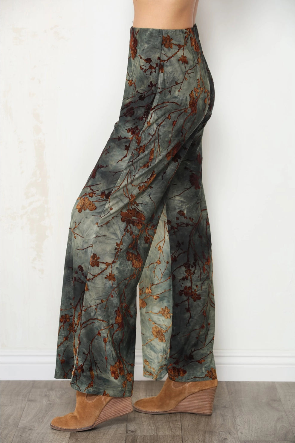 A person wearing wide-leg pants with a muted floral branch print in gray-green, brown, and rust tones, paired with tan wedge sandals, standing against a white wall.