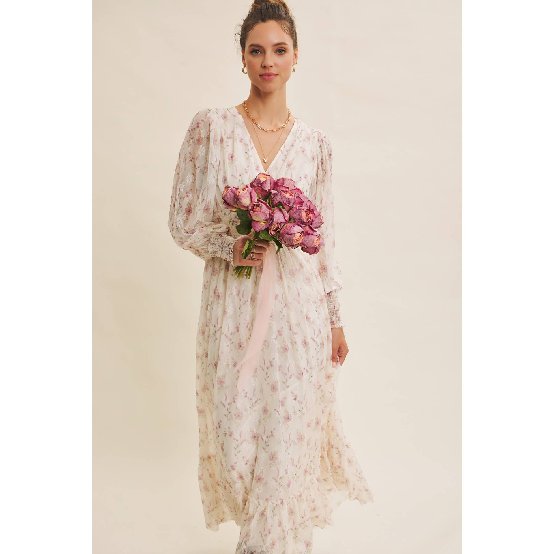 Front view — cream floral chiffon maxi dress with V-neck, sheer long sleeves, smocked waist, ruffle hem; model holding pink bouquet.