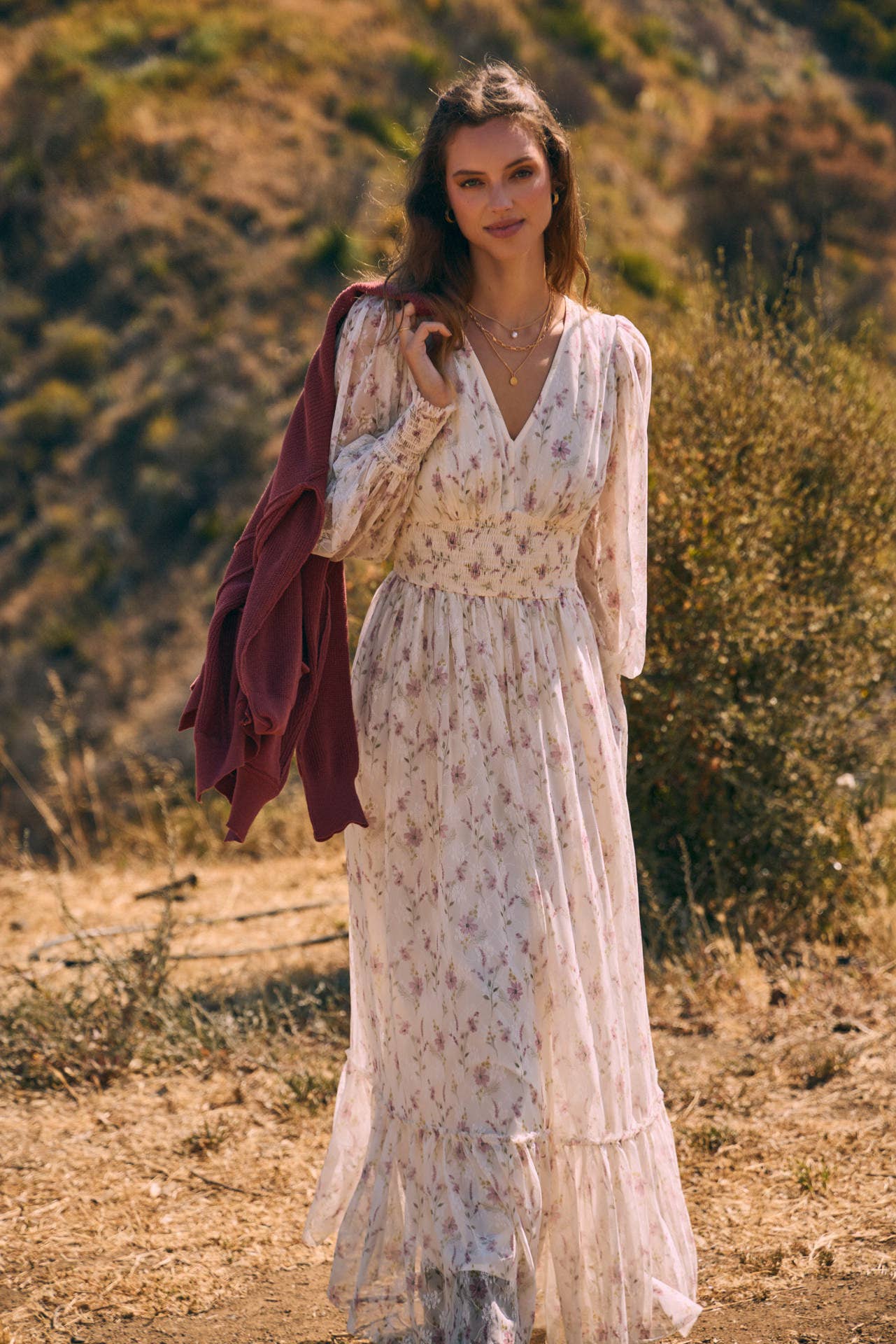 Front view — cream floral chiffon maxi dress with V-neck, sheer long sleeves, smocked waist, ruffle hem; model with mauve sweater over shoulder.