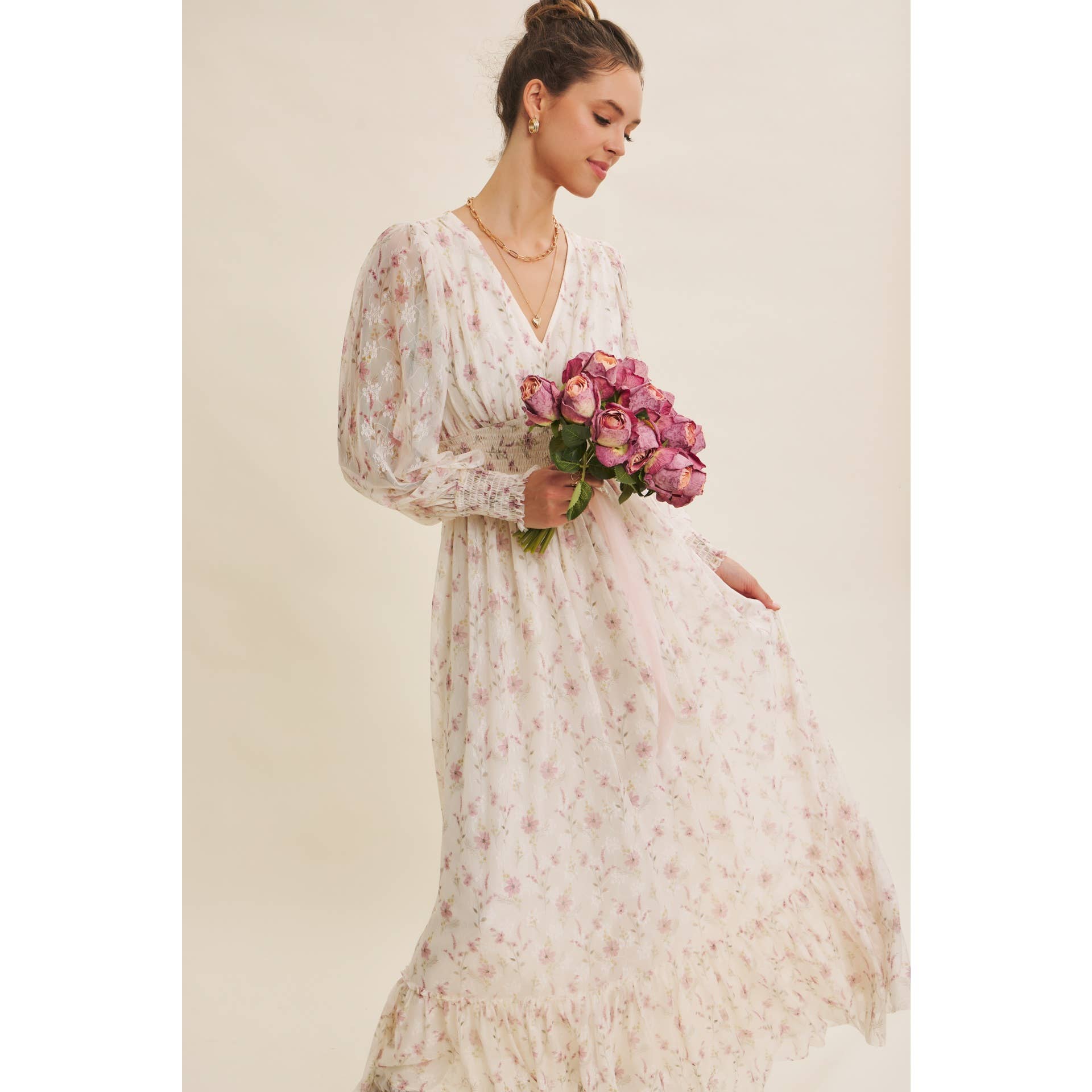 Front view — cream floral chiffon maxi dress with V-neck, sheer long sleeves, smocked waist, ruffle hem; model holding pink bouquet.