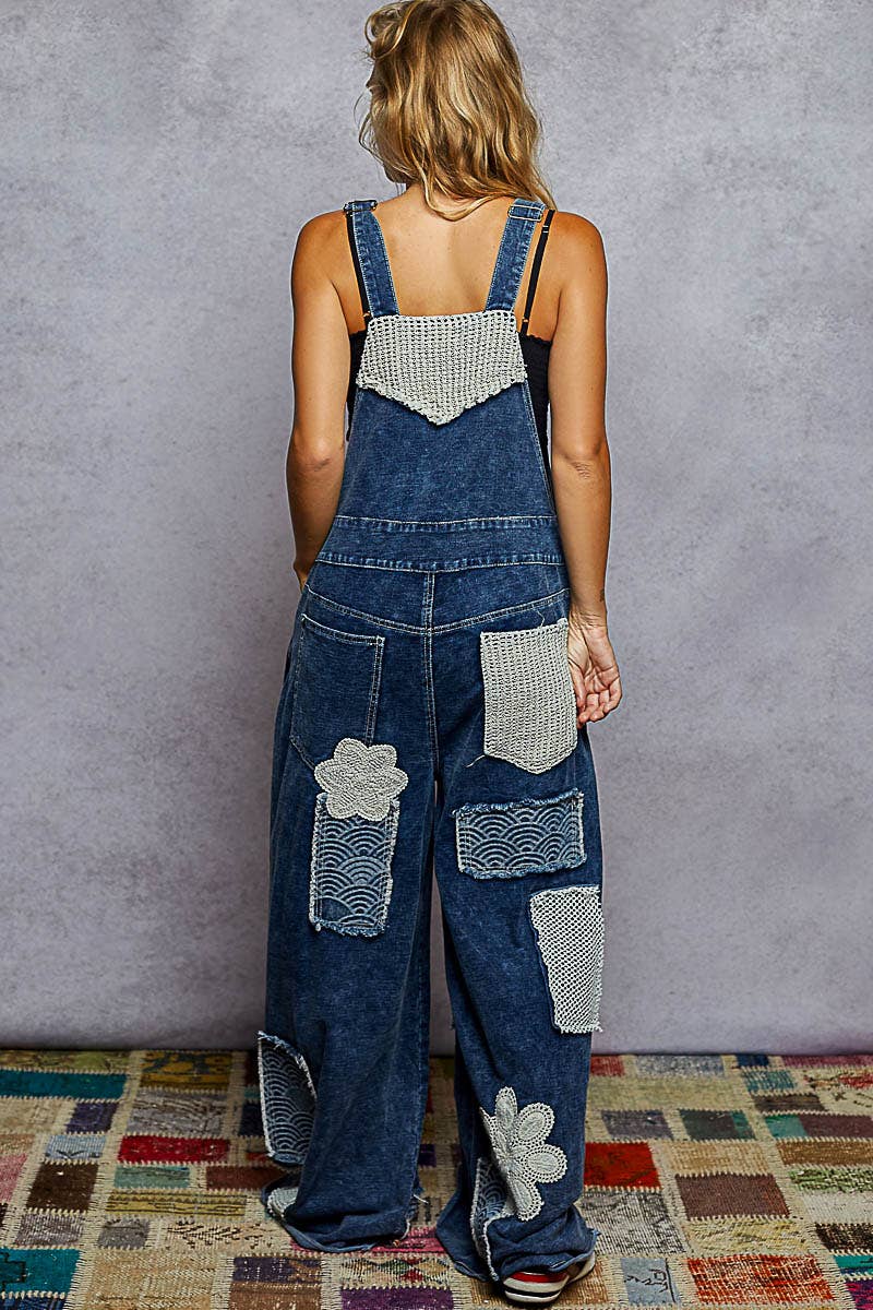 Back view of model wearing Boho denim patchwork overalls featuring wide legs, crochet lace appliqué patches, and vintage-inspired distressed detailing. Styled with a black crop top and sneakers for a relaxed, eclectic look.