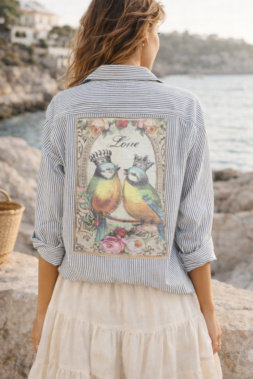 Woman wearing a jacket with a floral design and birds on the back, standing by a body of water.