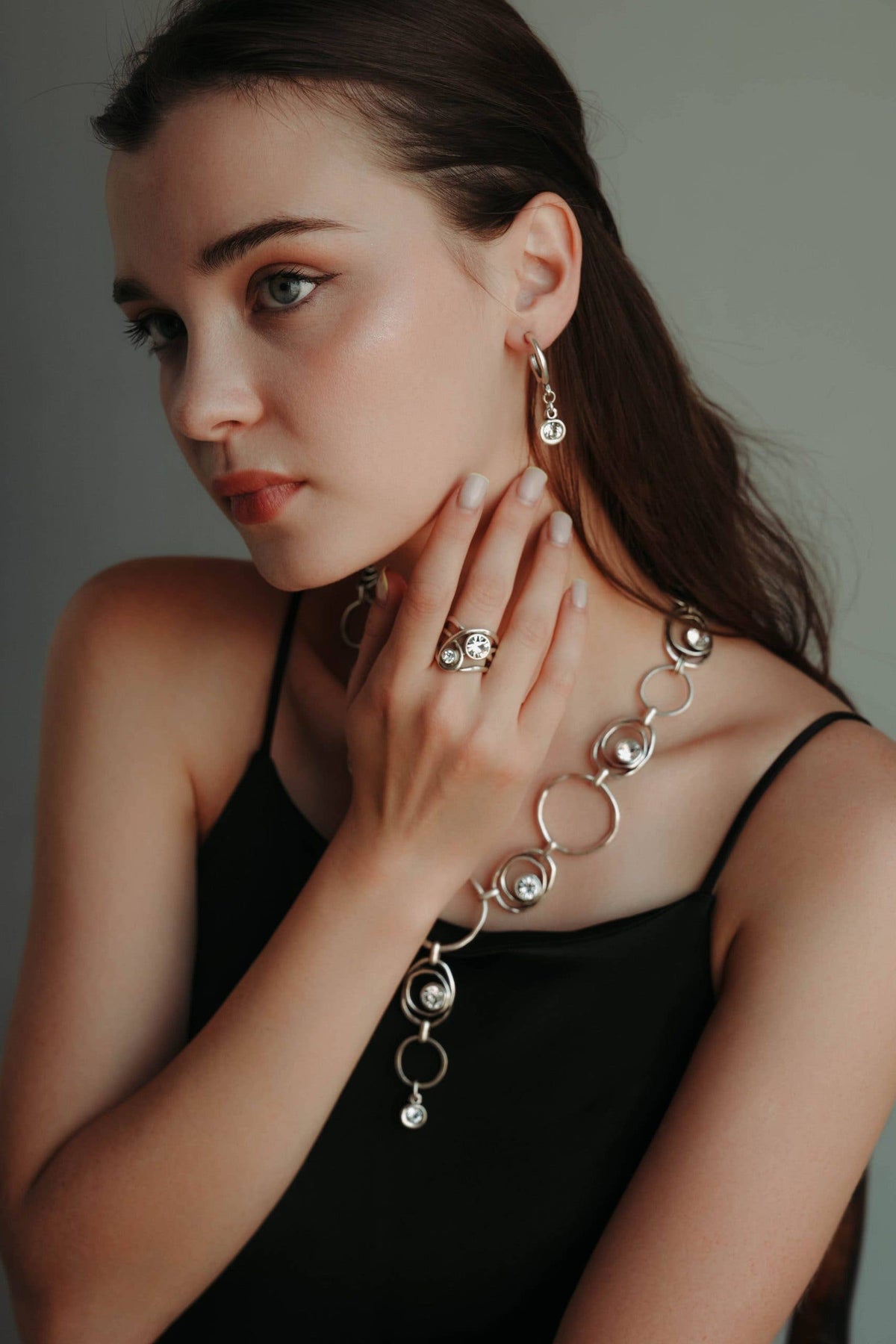 A woman wearing a silver-toned jewelry set featuring a link chain necklace, matching drop earrings, and a ring, styled with a black spaghetti-strap top against a neutral gray background.