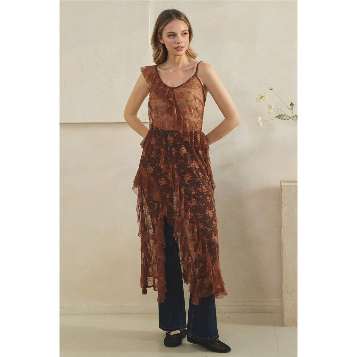 A woman models a copper sheer floral lace tunic top with ruffled layers over dark wide-leg pants, standing in a minimalist beige interior setting.